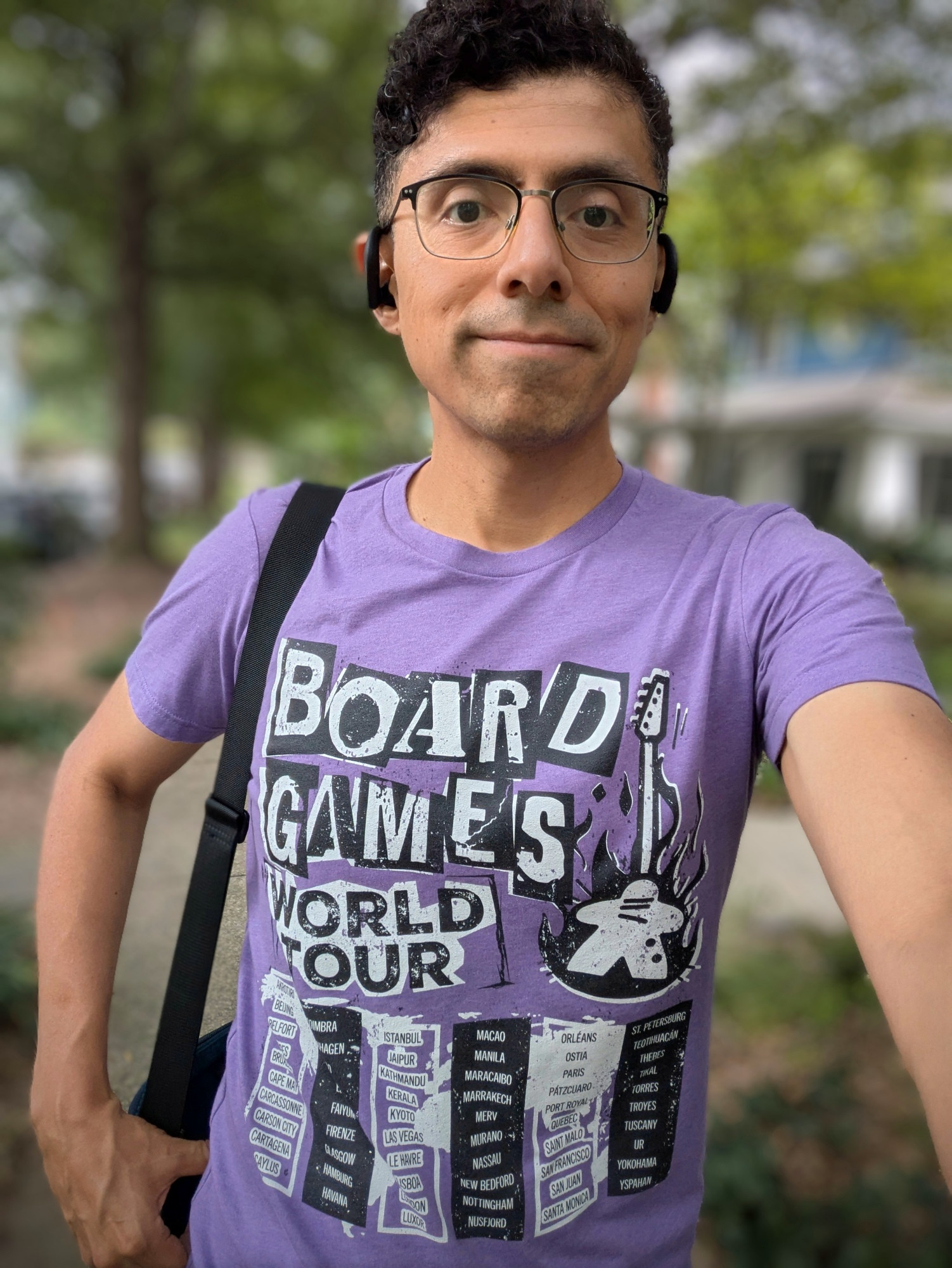 Daniel selfie wearing a "Board Games World Tour" shirt in a pink rock style, with a flaming meeple guitar and a list of cities.