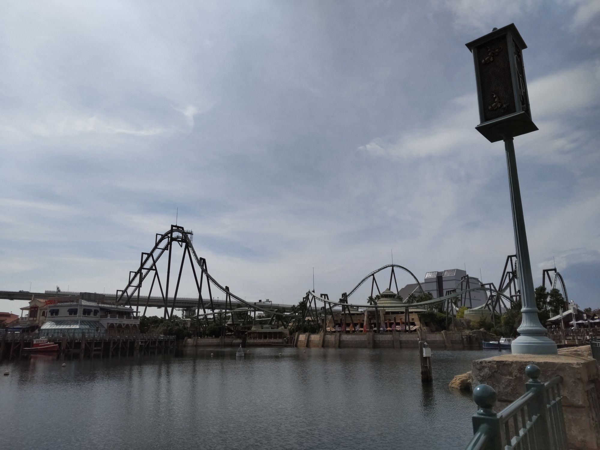 The lagoon at Universal Studios Japan looking across at the Jurassic Park area with the Flying Dinosaur roller coaster looming above it all, underneath a streaky, semi-blue sky.