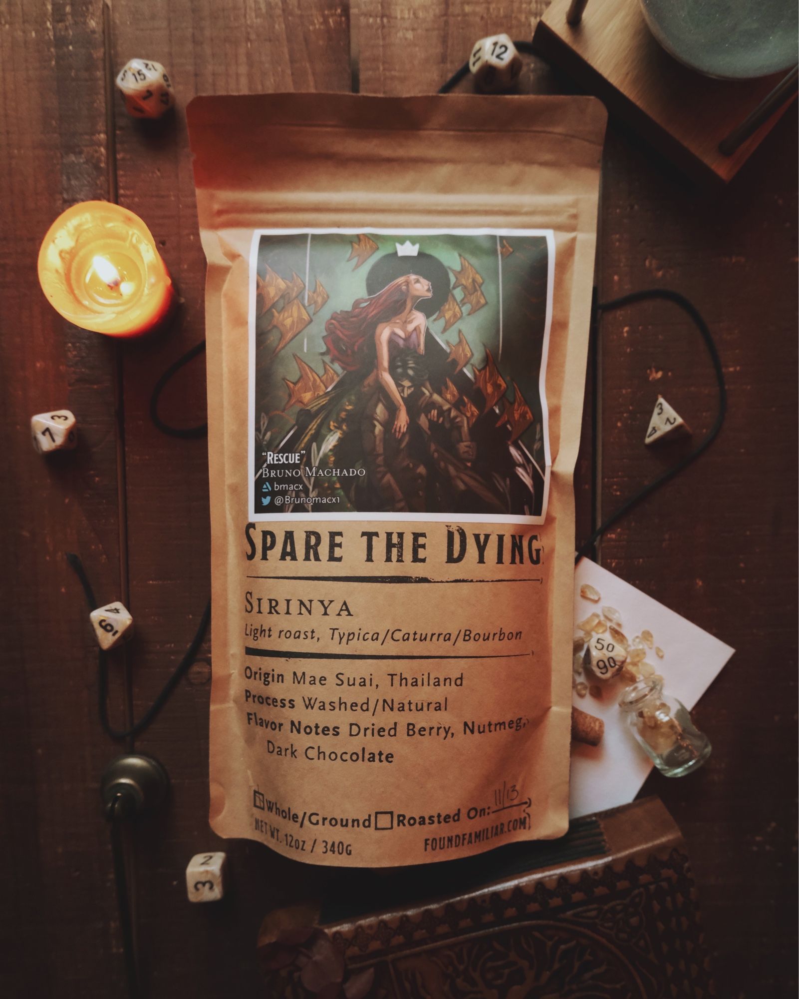 A kraft colored bag of Found Familiar Coffee laying on a wooden surface with dice and a candle framing it.  The title of the coffee is “Spare the dying” and the bag is adorned with artwork by Bruno Machado of a mermaid rescuing a drowning man.