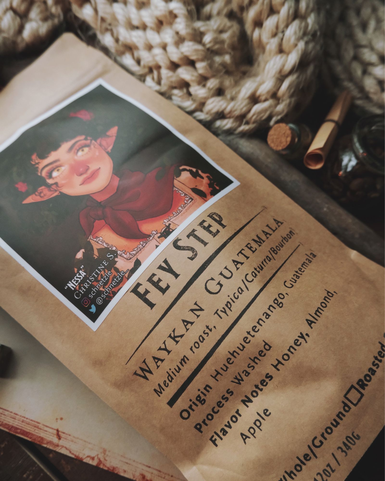 A bag of Found Familiar Coffee lies on an open book and framed by a thick burlap sack and various jars.  The bag is titled “Fey Step” and features artwork of an elven girl by ArcaneGold.