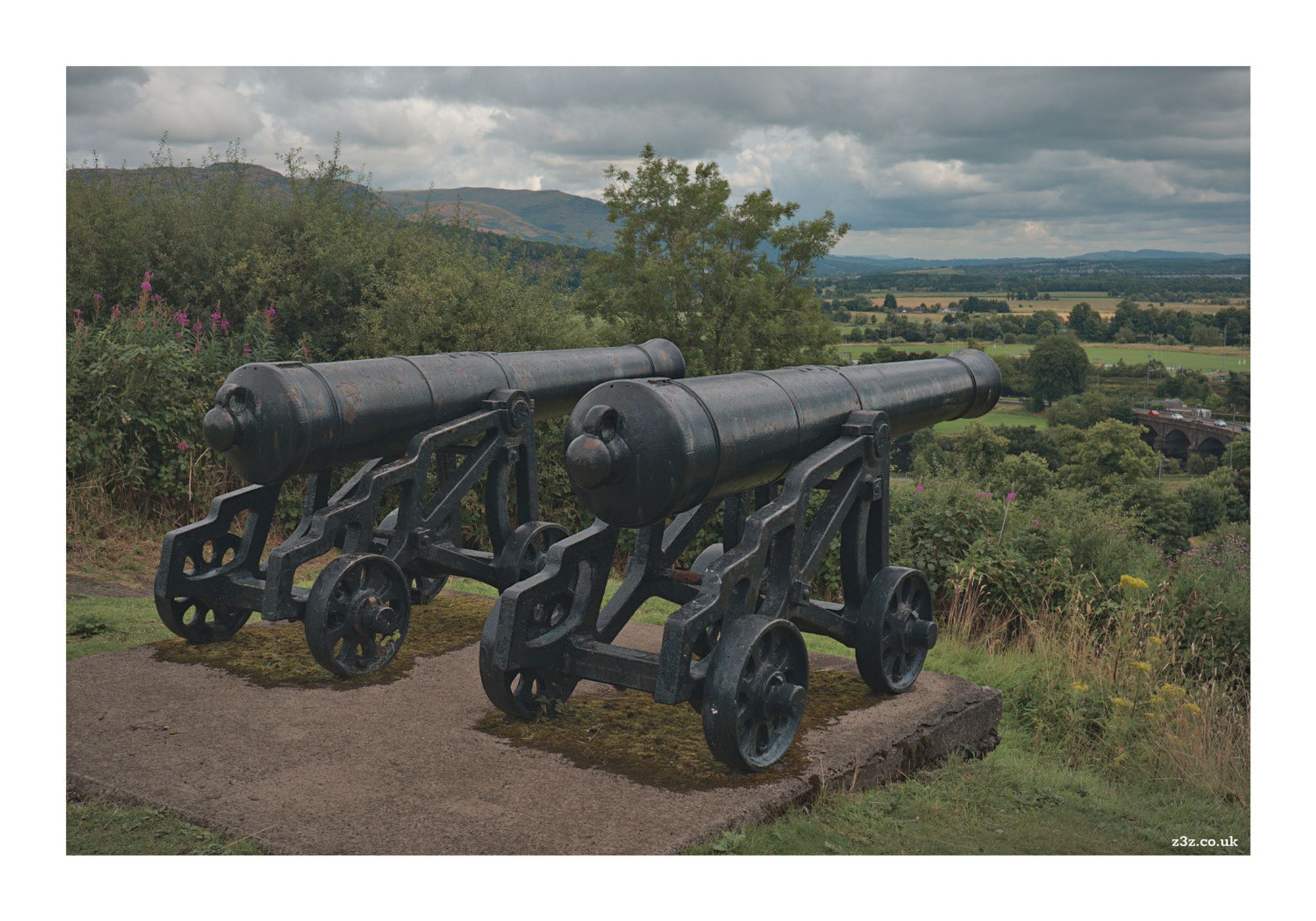 Two black cannons sitting side by side, and looking out over mostly rural countryside.