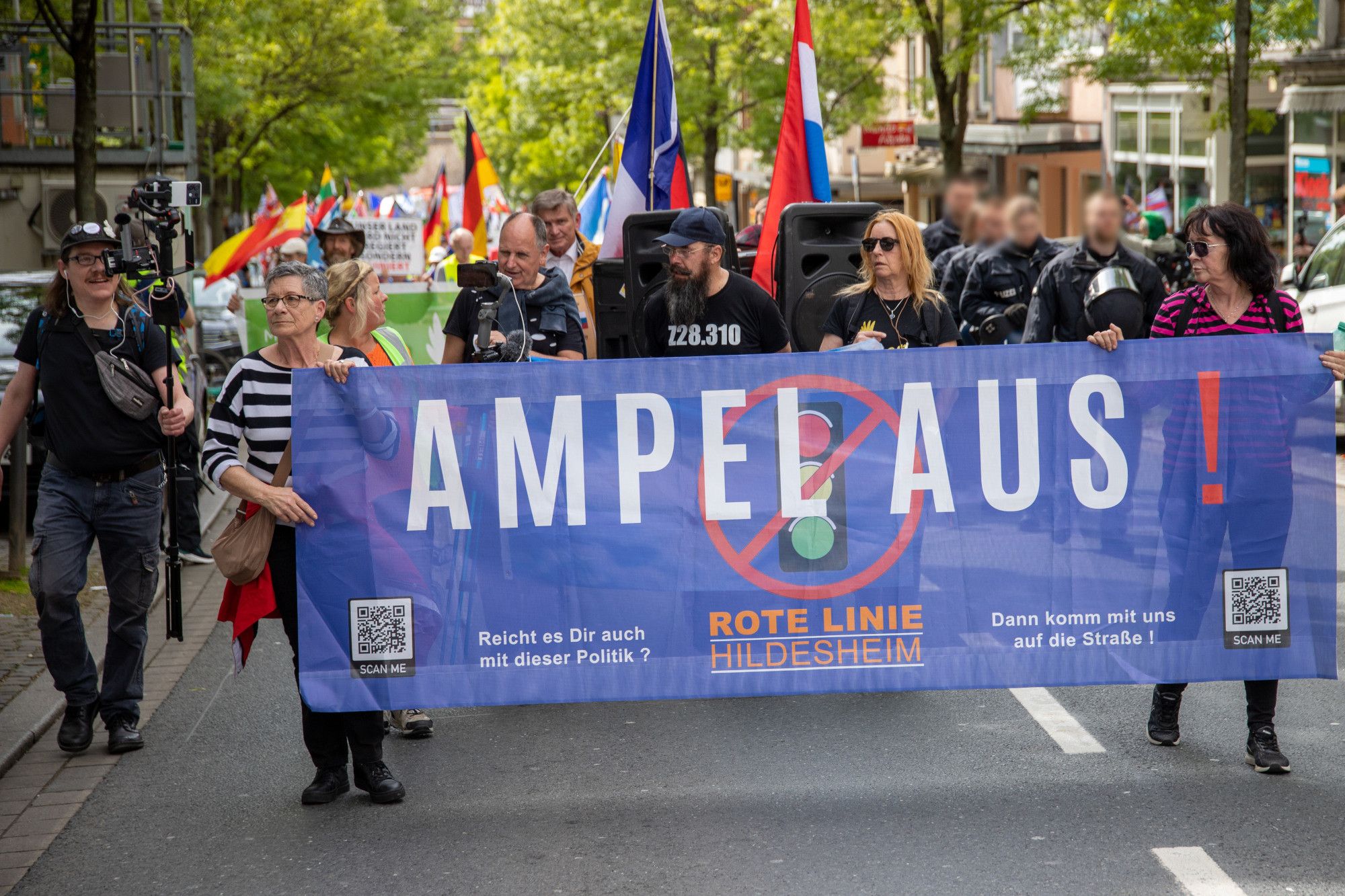 Fronttransparent des verschwörungsidiologischen Aufzugs:"Ampel aus! Rote Linie Hildesheim" auf blauem Grund mit einer durchgestrichenen Ampel im Hintergrund. Dahinter Menschen und Fahnen