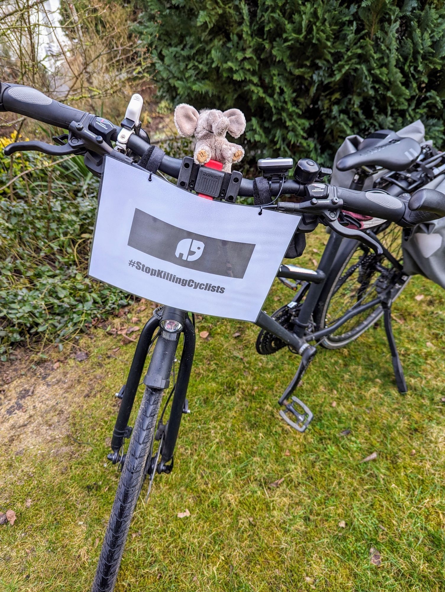 Schwarzes Fahrrad im Garten. Plüsch -Elefant sitzt auf Lenker. Am Lenker Schild befestigt, auf dem ein weißer Elefant auf schwarzem Untergrund zu sehen ist. Darunter steht Hashtag Stop Killing Cyclists