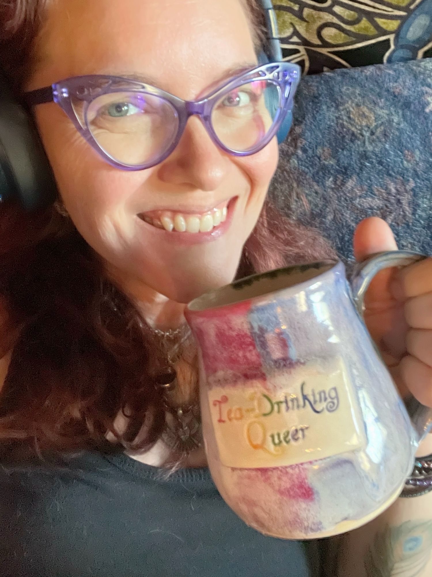 Sooj (white, femme, 44) sipping Dryad Tea from a Dryad Pottery mug labeled “Tea-Drinking Queer” in rainbow glaze, with curly rainbow script. She’s grinning at us over the lip of the cup, and her smile absolutely reaches her blue-green eyes, behind her purple cateye glasses. 