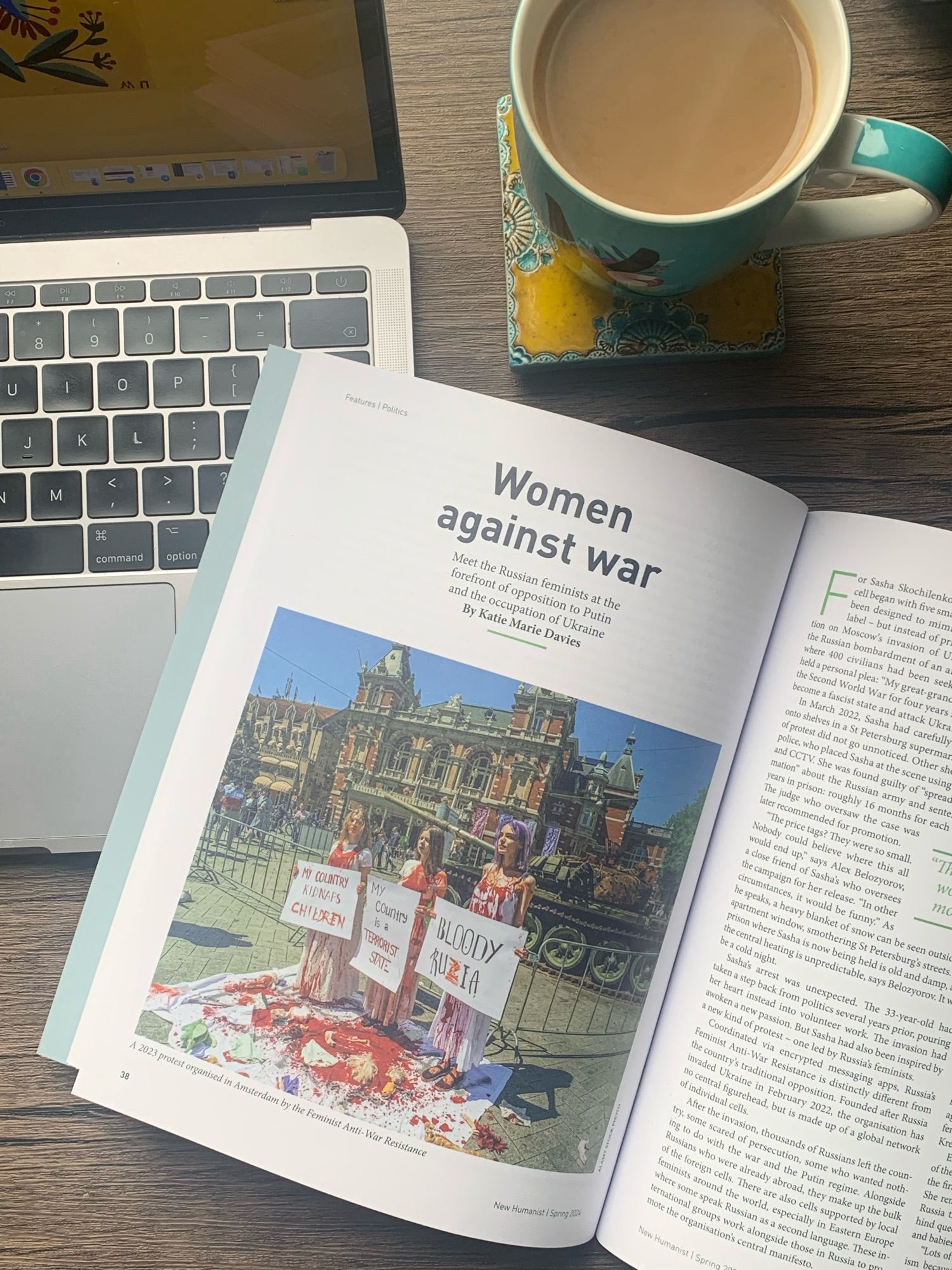 A picture of an open magazine next to a laptop and a cup of tea. The headline shown on the page of the magazine reads “Women against War”