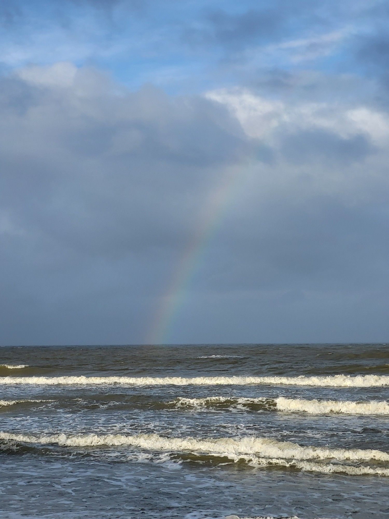 Ein halber Regenbogen - die linke Seite - über dem Meer mit leichtem Wellengang. Der Himmel ist dunkel bewölkt mit kleinen blauen Löchern.