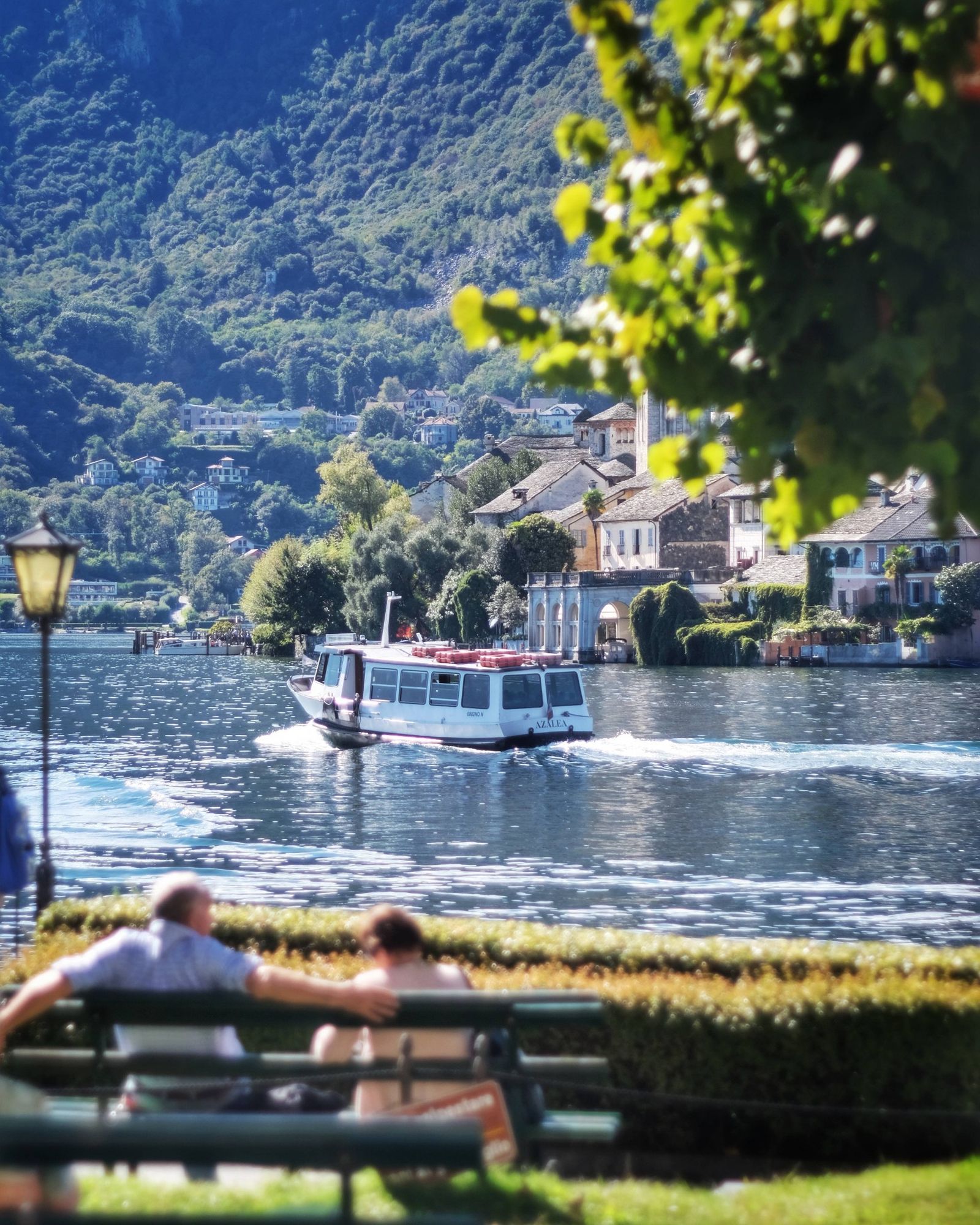 La barca sul lago,in primo piano due persone sedute su una panchina e in fondo le case su una isola in mezzo il lago