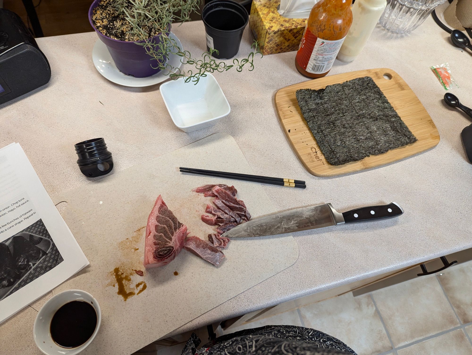 Counter of a cut of striated tuna, a bowl of soya sauce, chef's knife, cutting board with a sheet of nori, hot sauce, random rosemary plant.