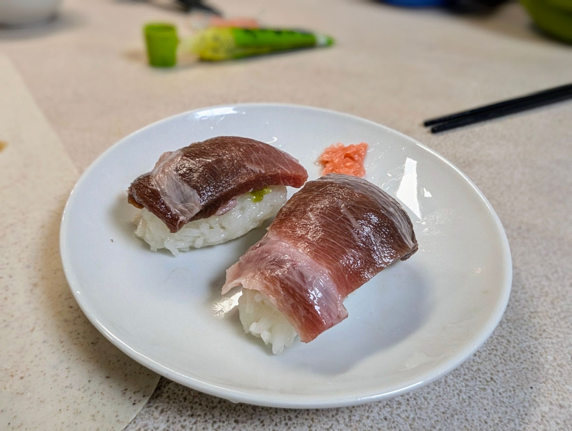 Two pieces of deep red and fatty tuna nigiri with pickled ginger on a small round plate, with pickled ginger, flanked by a tube of wasabi and the black chopsticks.