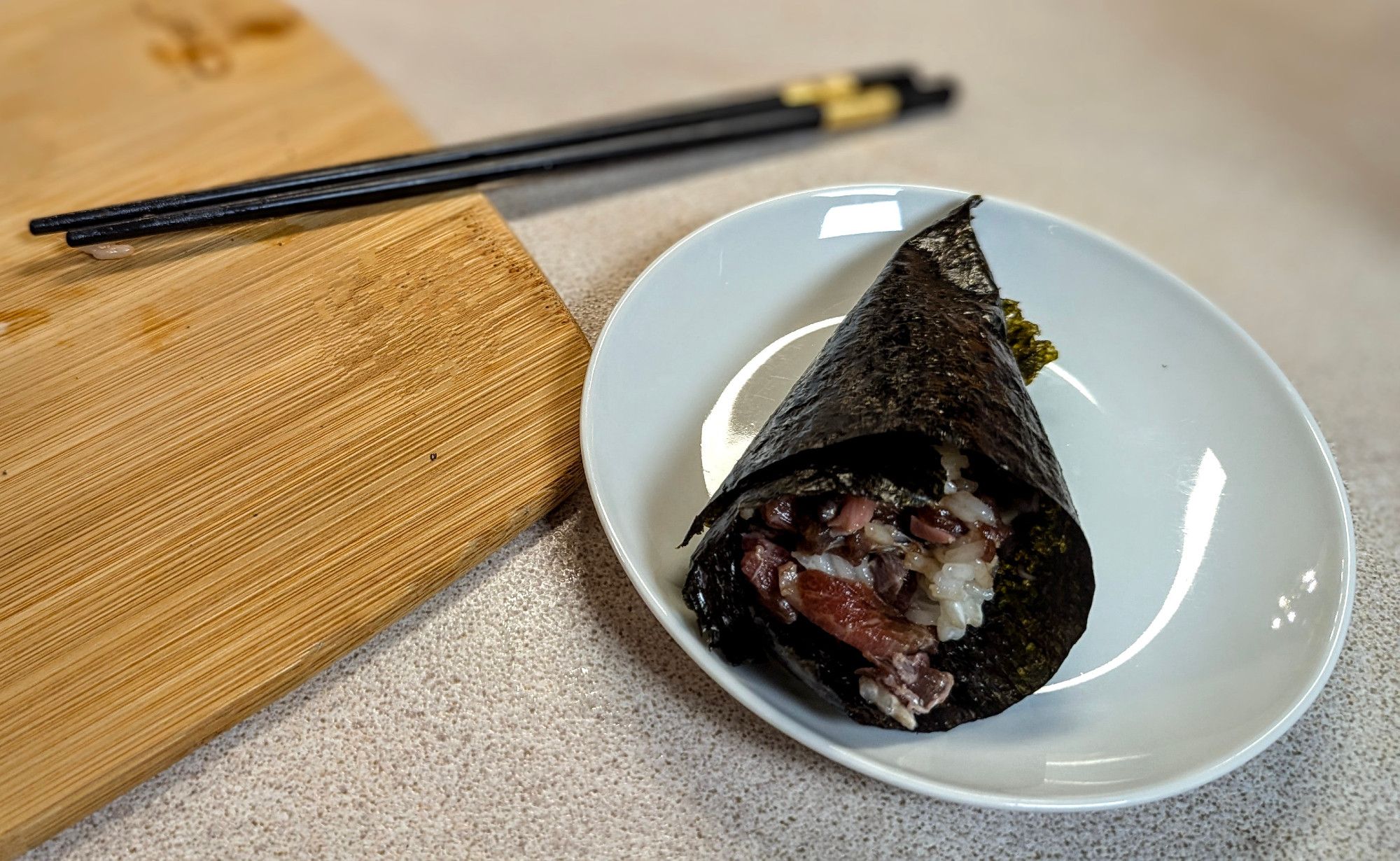 Close up of a reasonable spicy tuna hand roll on a round plate, with black chopsticks on a bamboo cutting board in the background.