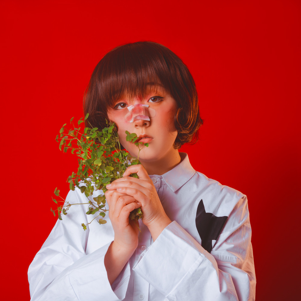 bright red background female character with a bloody bandaid in her nose holding a handful of clover leaves wearing a white shirt