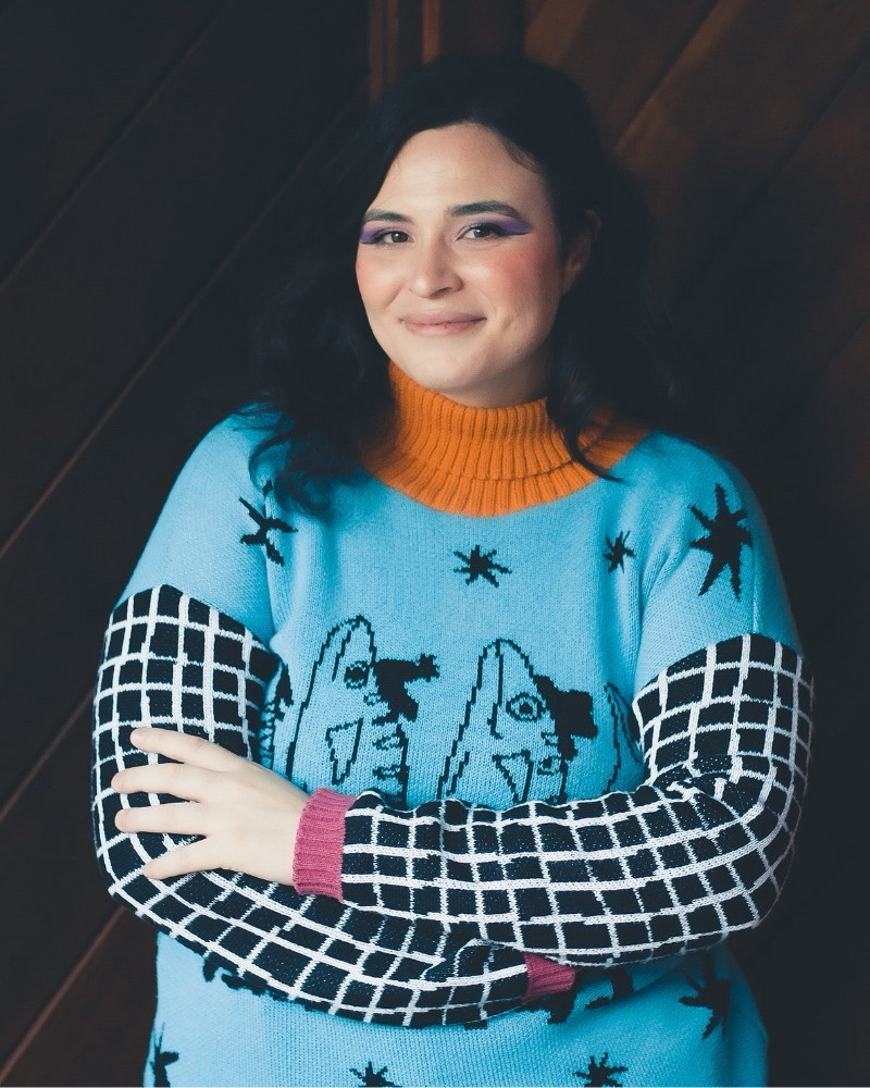 Foto. Fundo com ripas de madeira. Mulher branca de cabelo preto liso e ondulado abaixo dos ombros. Sombra roxa nos olhos. Sorriso leve no rosto. Está de braços cruzados. Usa uma blusa de frio no estilo suéter, todo colorido, a gola laranja, o tronco azul com desenhos pretos, estrelas espalhadas e dois peixes na vertical, em paralelo, as mangas com fundo preto e listras brancas na vertical e na horizontal, o punho rosa.