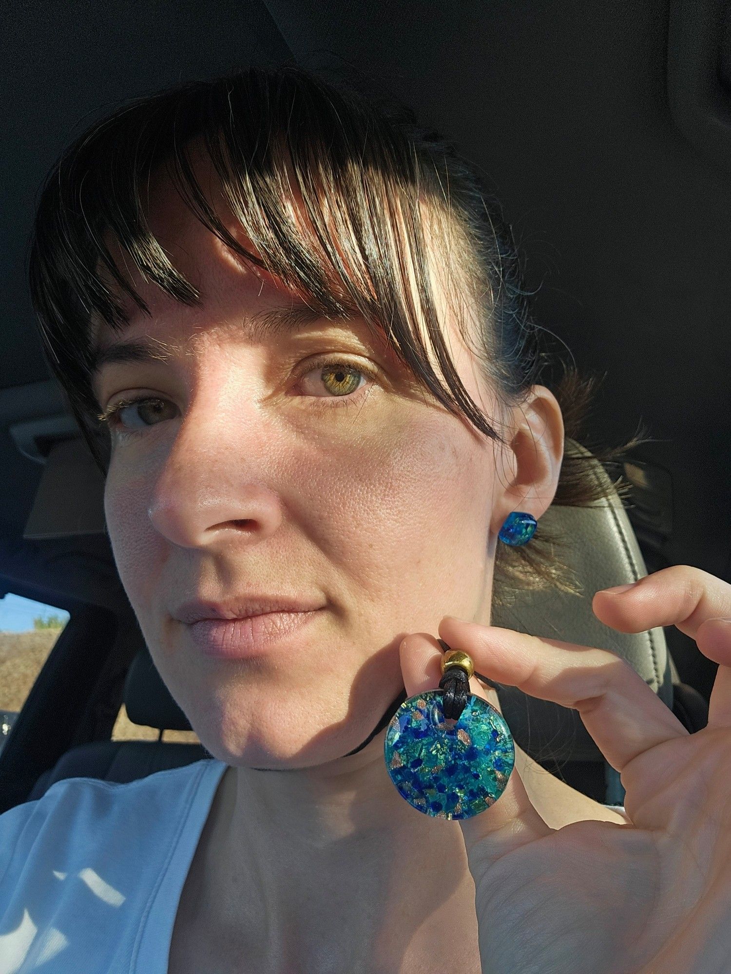 Selfie in a car. Me showing my necklace and matching earrings. The necklace is round, about an inch in diameter, and full of multicolor sparkles. The predominant color is turquoise. The earrings are maybe a third of an inch square in the same glass material.