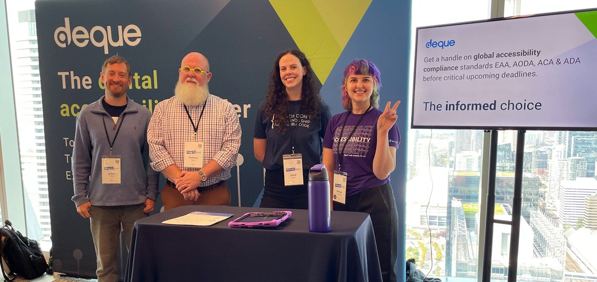 Deque employees Josh, Matthew, Grace and Rachael smiling together in front of the Deque booth with the downtown Toronto cityscape in the backdrop.