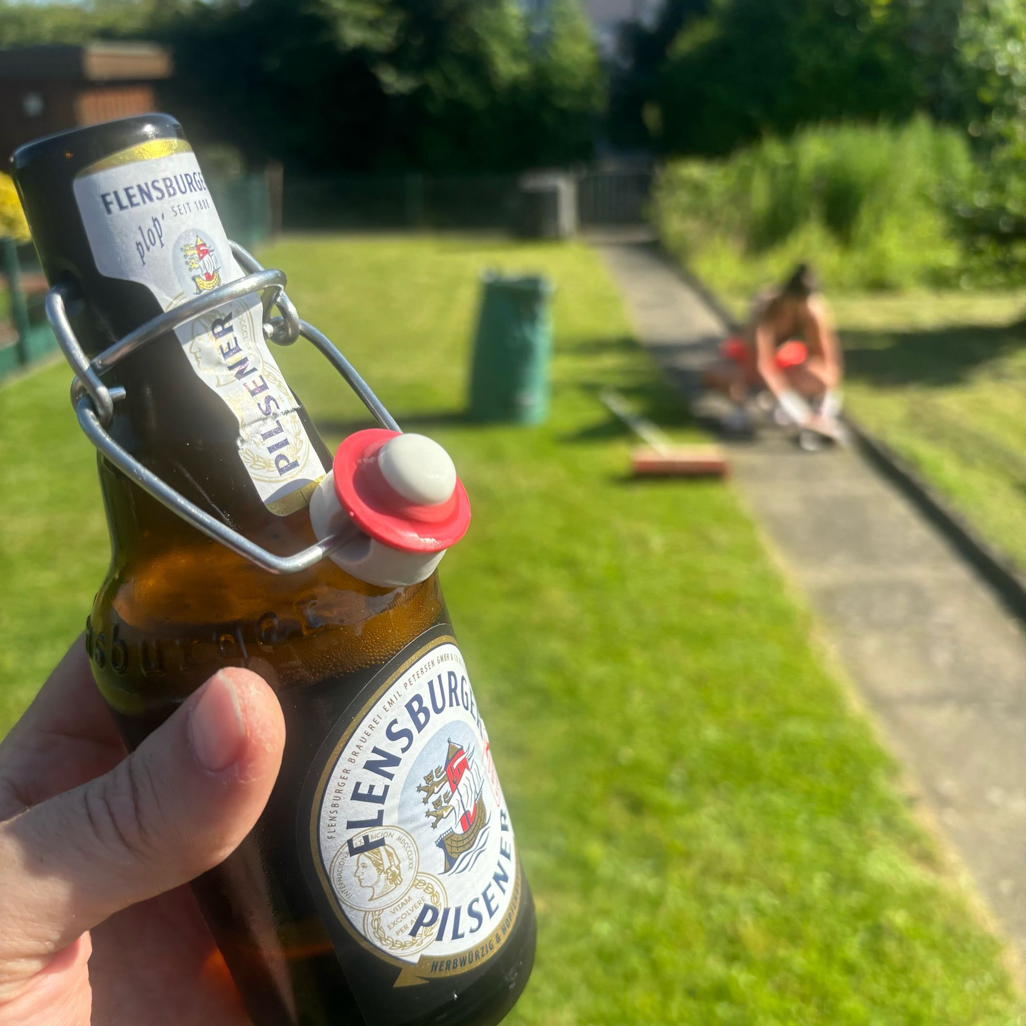 Eine Buddel Flens, im Hintergrund die Holde beim Fegen im Garten