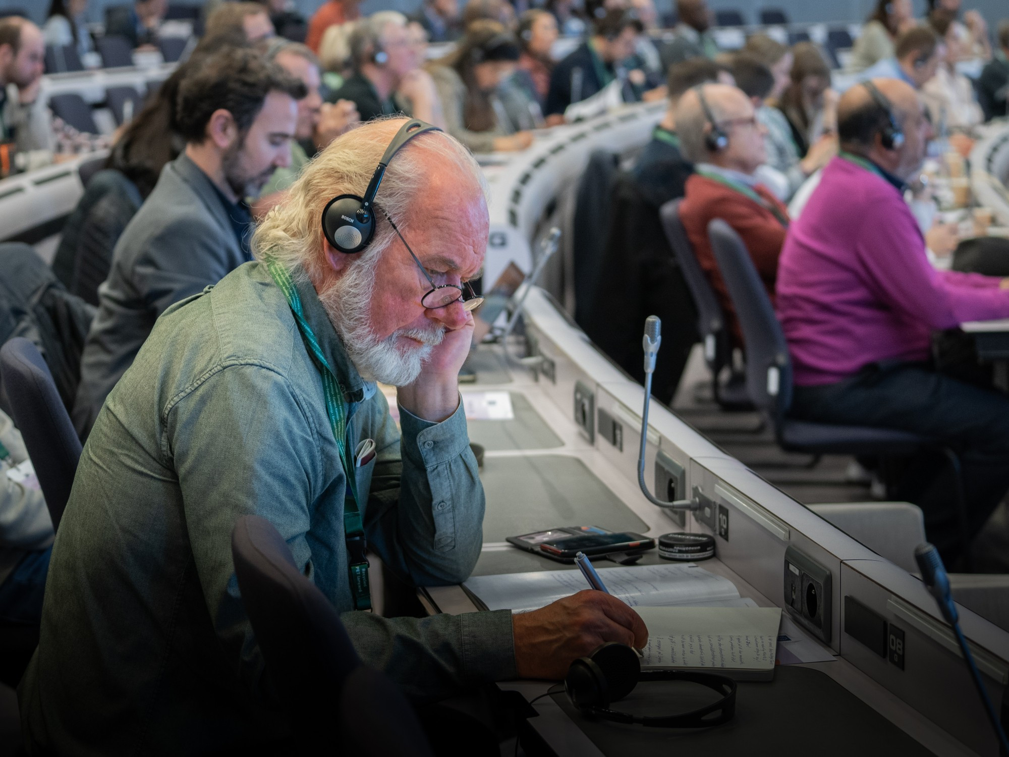 A photo of a person taking notes while listening to the headphones in a room filled with people.