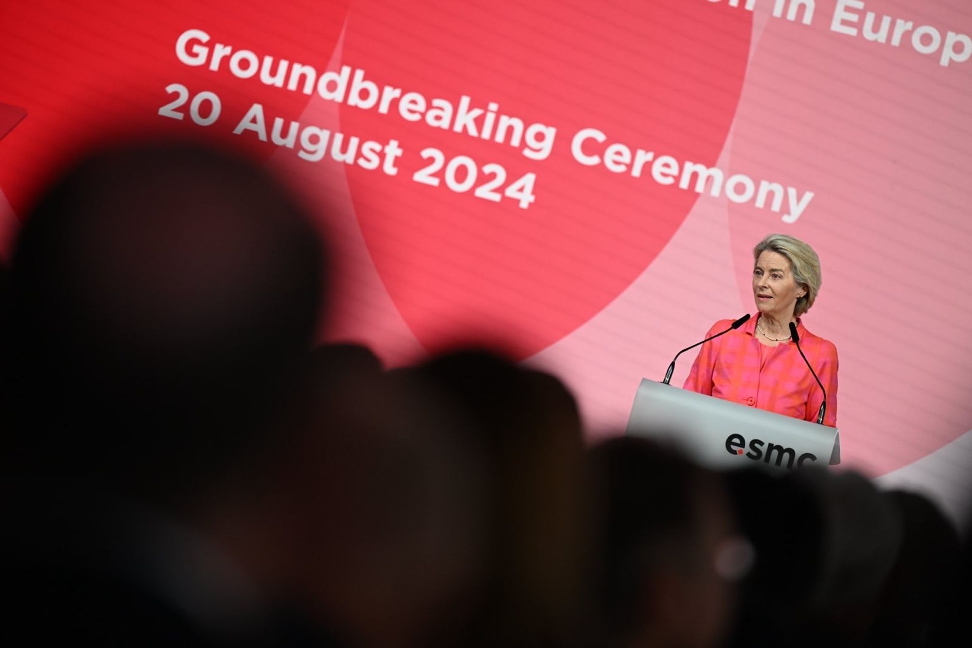 The image shows Ursula von der Leyen, President of the European Commission, speaking at the Ceremony of ESMC on August 20, 2024. She is standing at a podium with the event's branding visible behind her on a large red screen. Von der Leyen is dressed in a pink jacket and is addressing the audience.