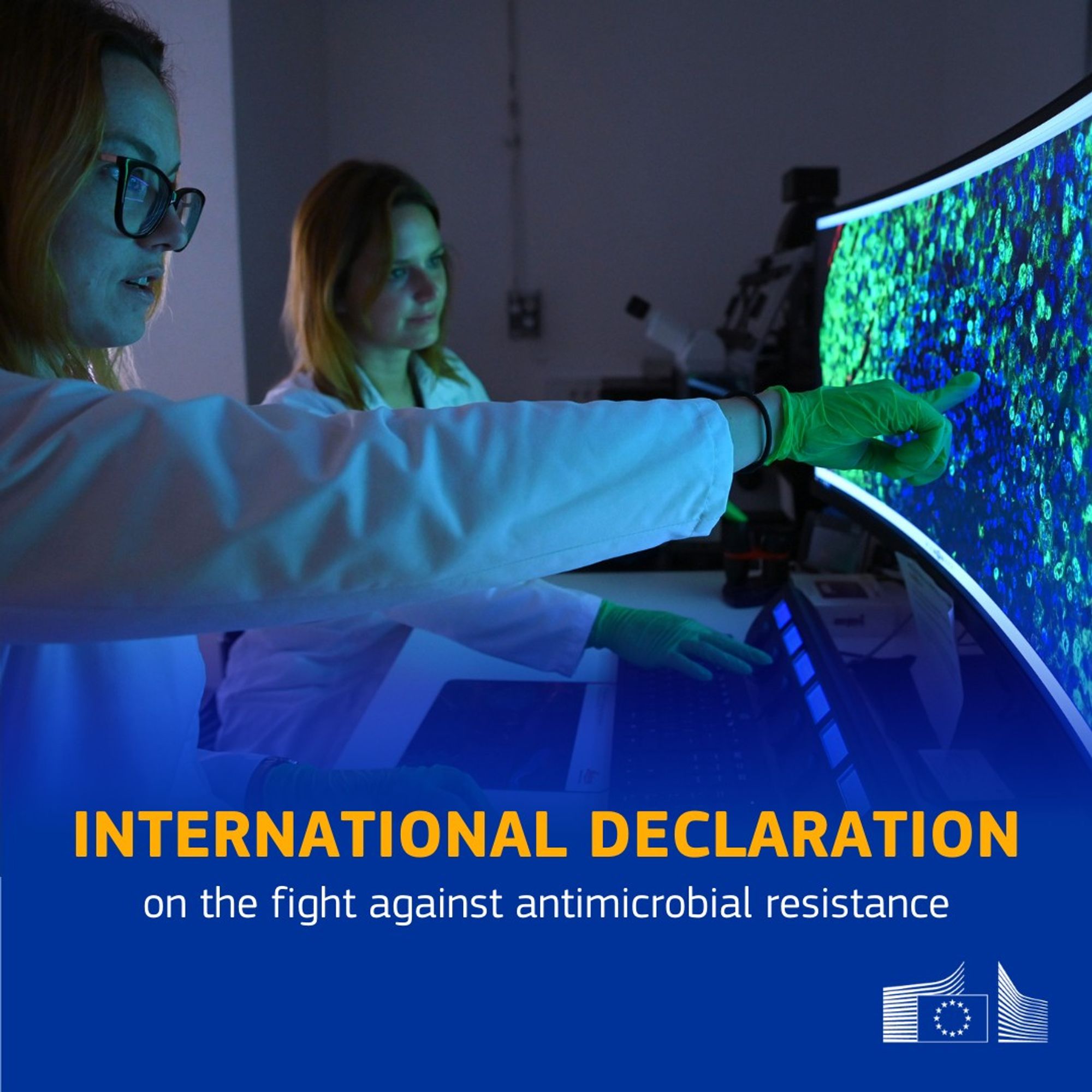 Two scientists, wearing lab coats and green gloves, are analyzing data on a large, curved computer screen displaying microscopic imagery in shades of blue and green. The text overlay reads "International Declaration on the fight against antimicrobial resistance" with the European Commission logo in the bottom right corner. The scene takes place in a dimly lit laboratory, emphasizing the work being done in scientific research and healthcare.