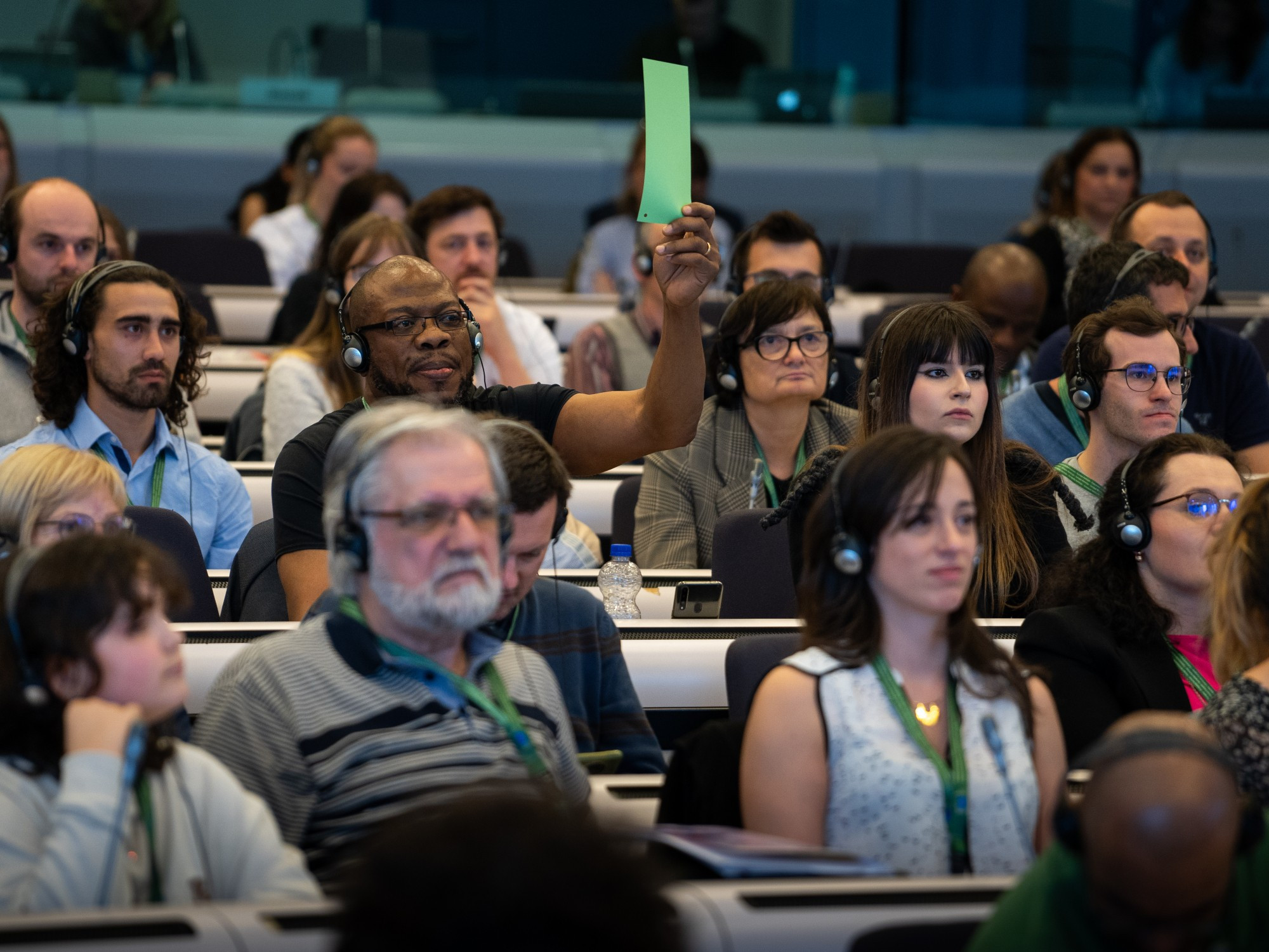 A group of people in a conference room listening to someone speaking while a person in the midst of them holds and wave a piece of paper.