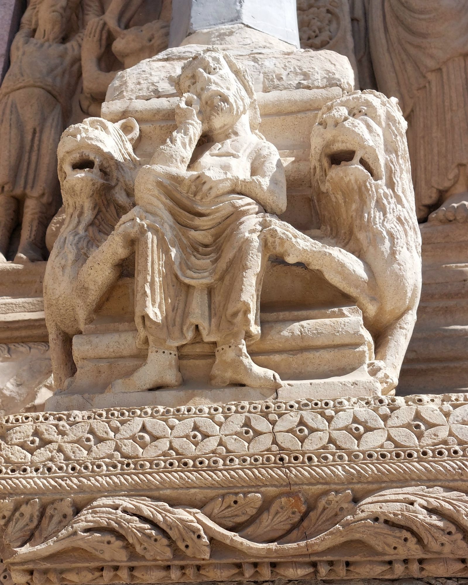 Detail of a sculpture on a Churchill Portal in Arles, France, depicting a sad-faced monk sitting with his head leaning on his right hand. Two lions on each side of him are putting one of their paws on his knees. They also look somewhat disappointed.