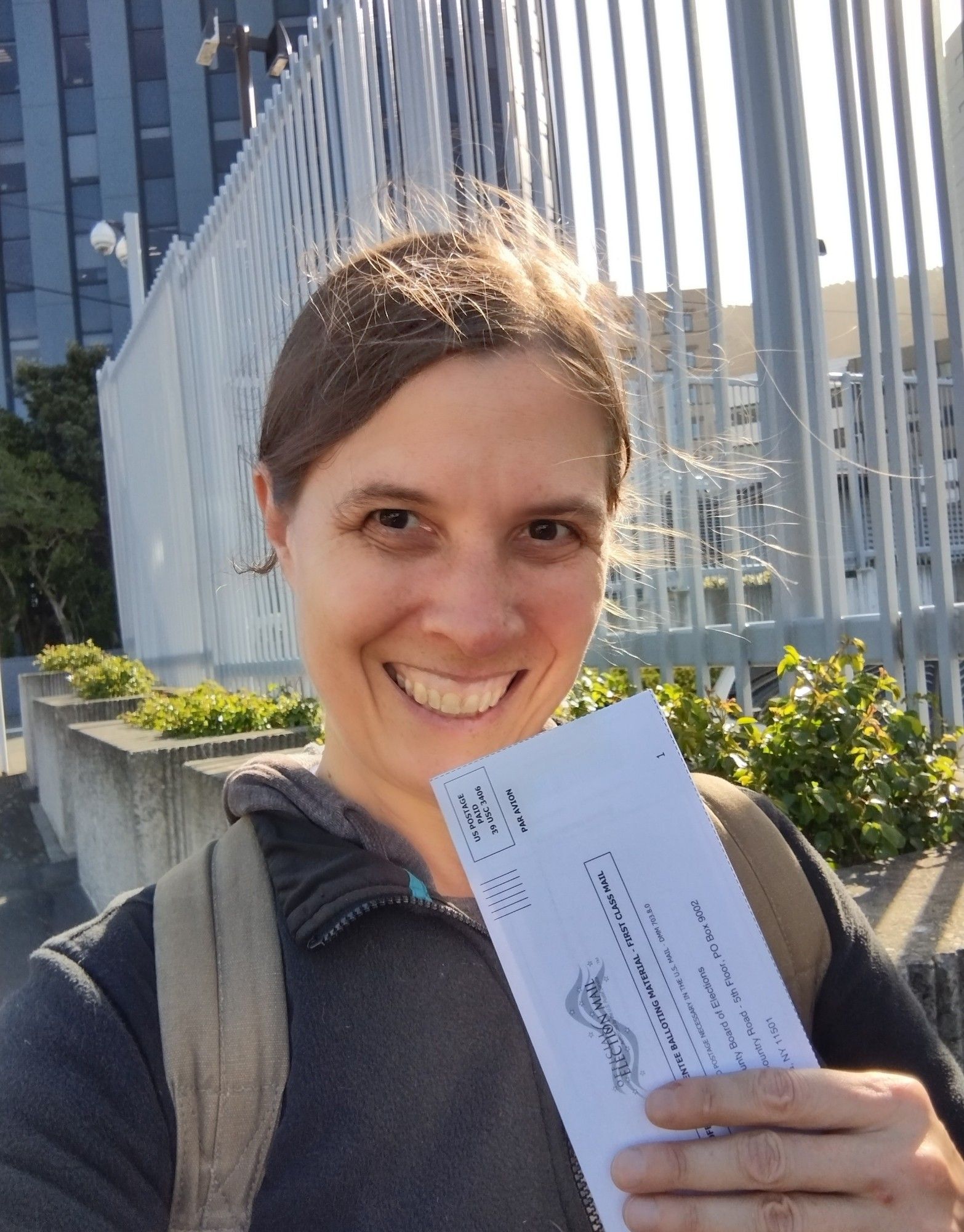 A photo of me smiling and holding my absentee ballot envelope outside the US embassy. It's windy as usual, and my hair is escaping my clip and blowing all over the place.