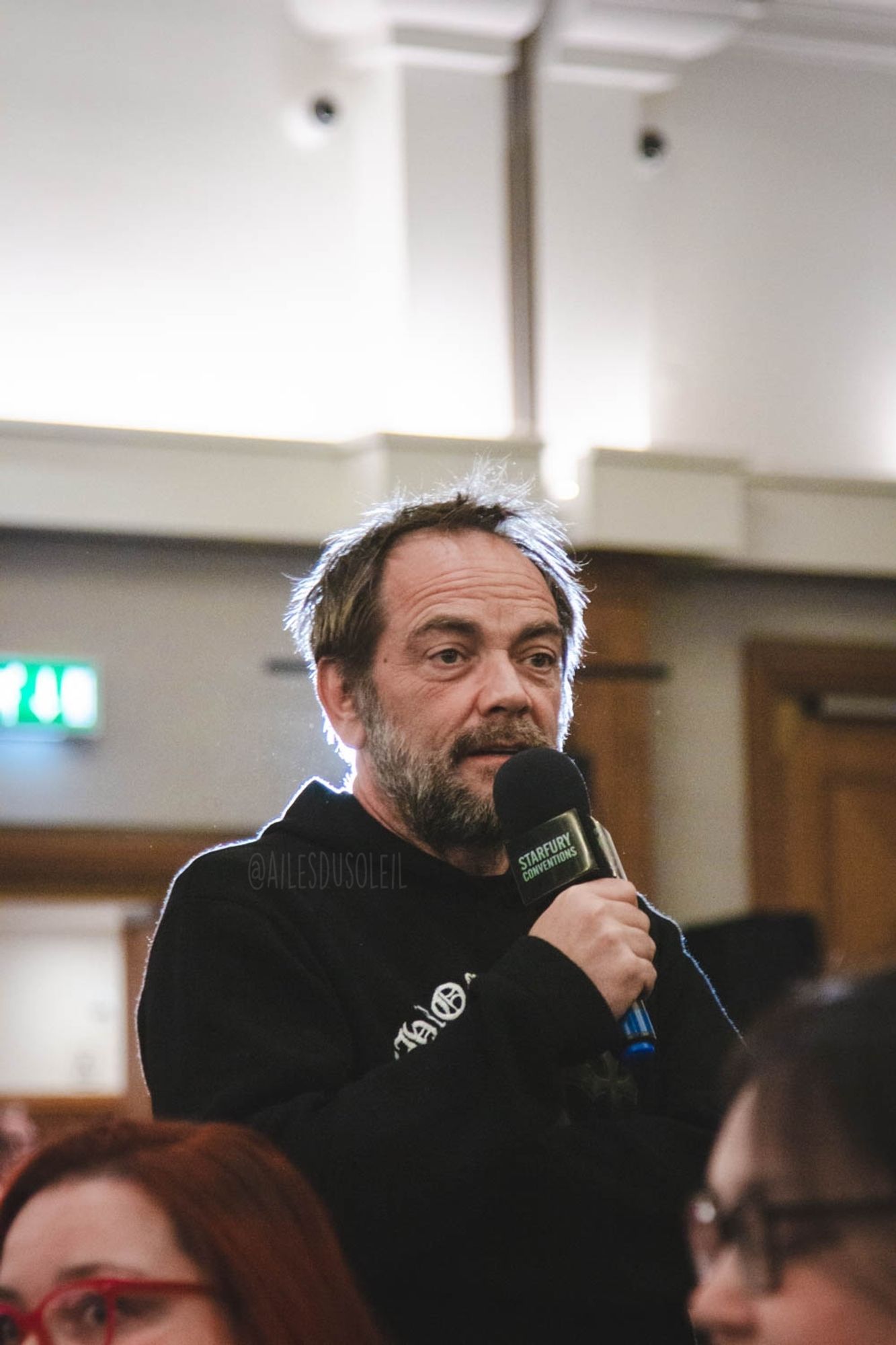 Mark is walking between the crowd. It is a closeup and he is visible midsection up. There is a lamp behind him making him have a white outline around his head. He is turned slightly towards the right side of the photo while talking into the microphone and looking at a person who asked a question. He is wearing a black pullover hoodie with white gothic text on it.