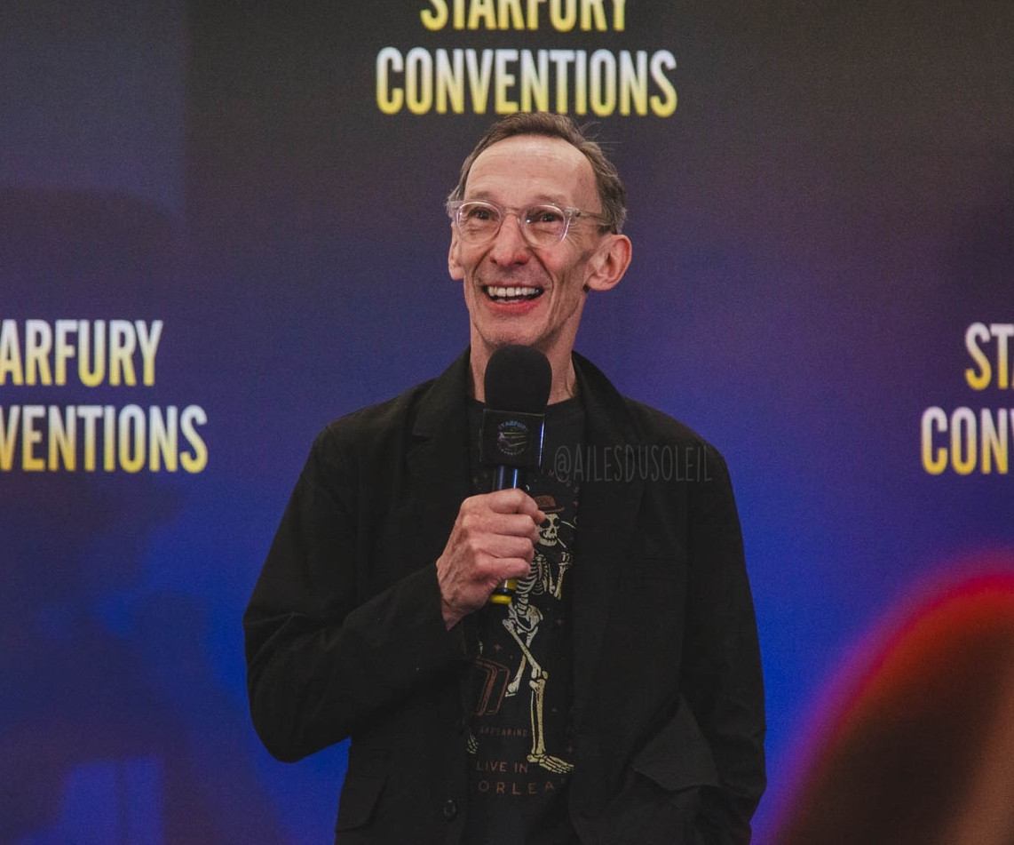 Julian is standing on stage visible from belt up. His body is turned towards the camera, but his face and his eyes are turned slightly left. He is smiling while listening to a person asking a question. He is holding his microphone chin high in his right hand while his right hand is in his right pants pocket. He is wearing a black t-shirt with a skeleton on it and a black blazer. He has see-through rimmed glasses.
