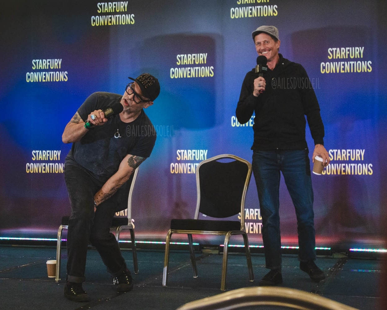 They are standing on stage, Todd on the left side of the photo and Corin on the right. Both are visible fully. Todd is somewhat bent to the right side of the photo sideways from the middle. Both his hands are bent together from the knees and his left arm is straight down with his hand between his thighs. He is speaking into the microphone in his right hand while looking towards the right side off frame. Nemec is standing up on the right side mid movement leaning on his right leg. He is smiling into his microphone that is mouth high in his right hand. His left arm is relaxed next to him holding a coffee cup. 

Todd is wearing black sneakers, jeans, black t-shirt and a black baseball cap. He has black framed glasses, some tattoos, wears multiple rings and a ring shaped necklace. 

Corin is wearing black shoes, blue jeans, black pullover and a gray beanie type of hat. He has a moustache and is wearing a round silver like ring on his right ring finger.