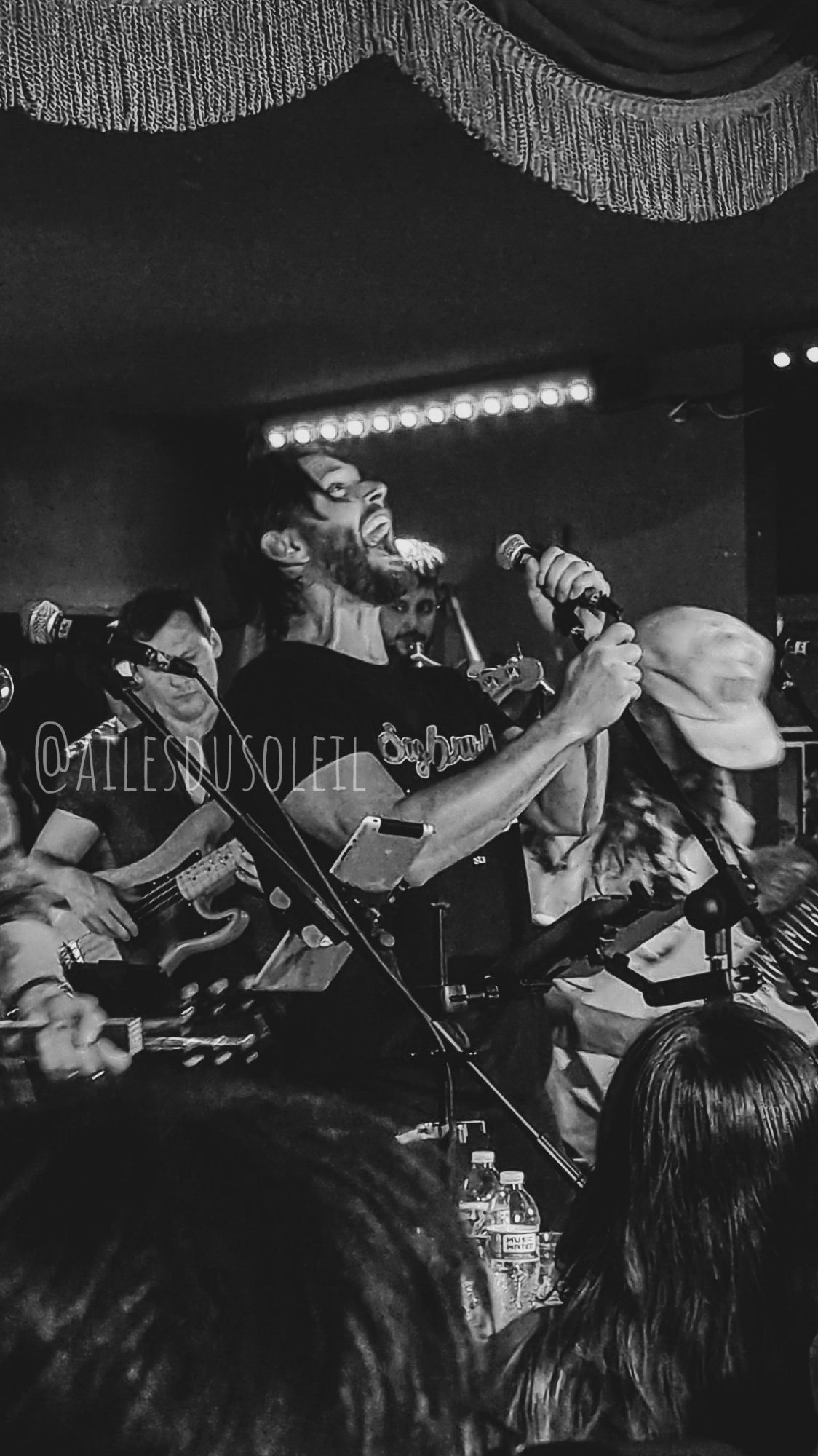 Black and white photo of Jensen Ackles on stage belting strong notes into the microphone which he is holding with both of his hands. He is slightly tilted back, mouth wide open and veins visible on his neck.