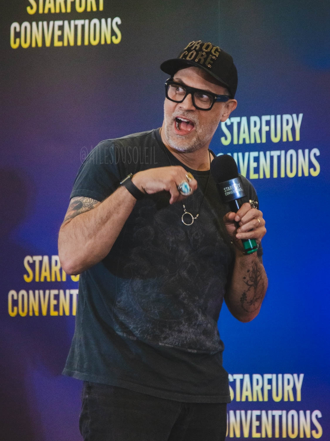 Todd is standing on stage, body facing about ⅔ to the right of the photo while his head is turned towards the left of the photo and he is visible from upper thigh up. He is making a cat like pose with his arms. They are bent against the front sides of his chest, fingers curled forward. He has a not really shocked face on - mouth open big and eyes big. His microphone is in his left hand collarbone height. He is wearing black jeans, black t-shirt and a black baseball cap. He has black framed glasses, some tattoos, wears multiple rings and a ring shaped necklace.