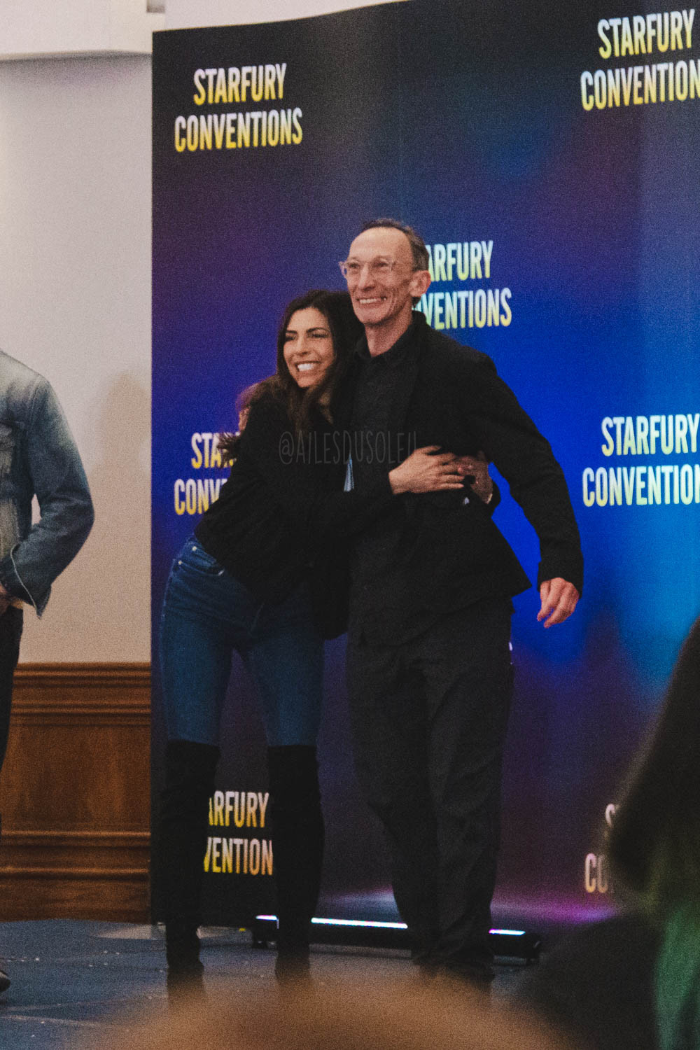 Cindy (left) and Julian (right) are standing on stage, Cindy bent forward hugging him with arms around his torso. Julians right arm is around Cindy on her upper back and the other arm is on his left side a bit further away. Cindy is wearing black over knee boots, dark jeans and a black jacket. Julian is wearing all black - pants, shirt and jacket. Both are smiling big