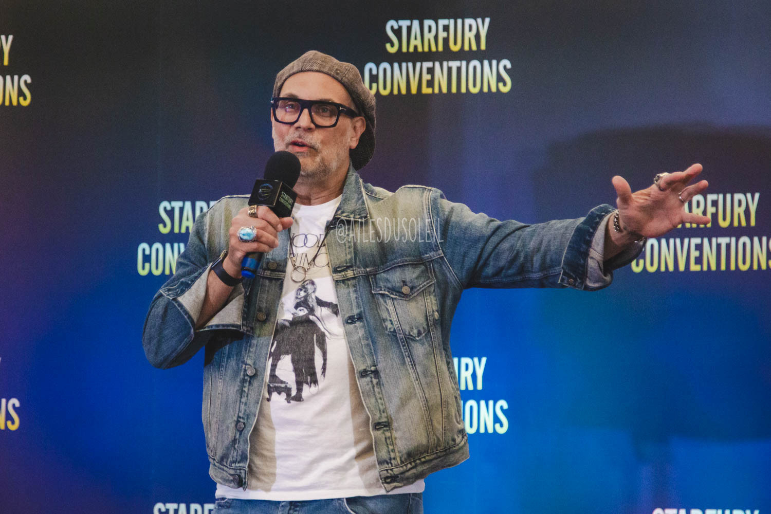 the camera. He is looking in the same direction. He is speaking into his microphone which he is holding with his right hand to his mouth. His left arm is stretched out to his side a bit mid motion explaining something. He is wearing blue jeans, white t-shirt, a discoloured light blue jean jacket and a backwards newsboy cap. He has black rimmed glasses on and is wearing multiple rings on multiple fingers and a ring shaped necklace. 