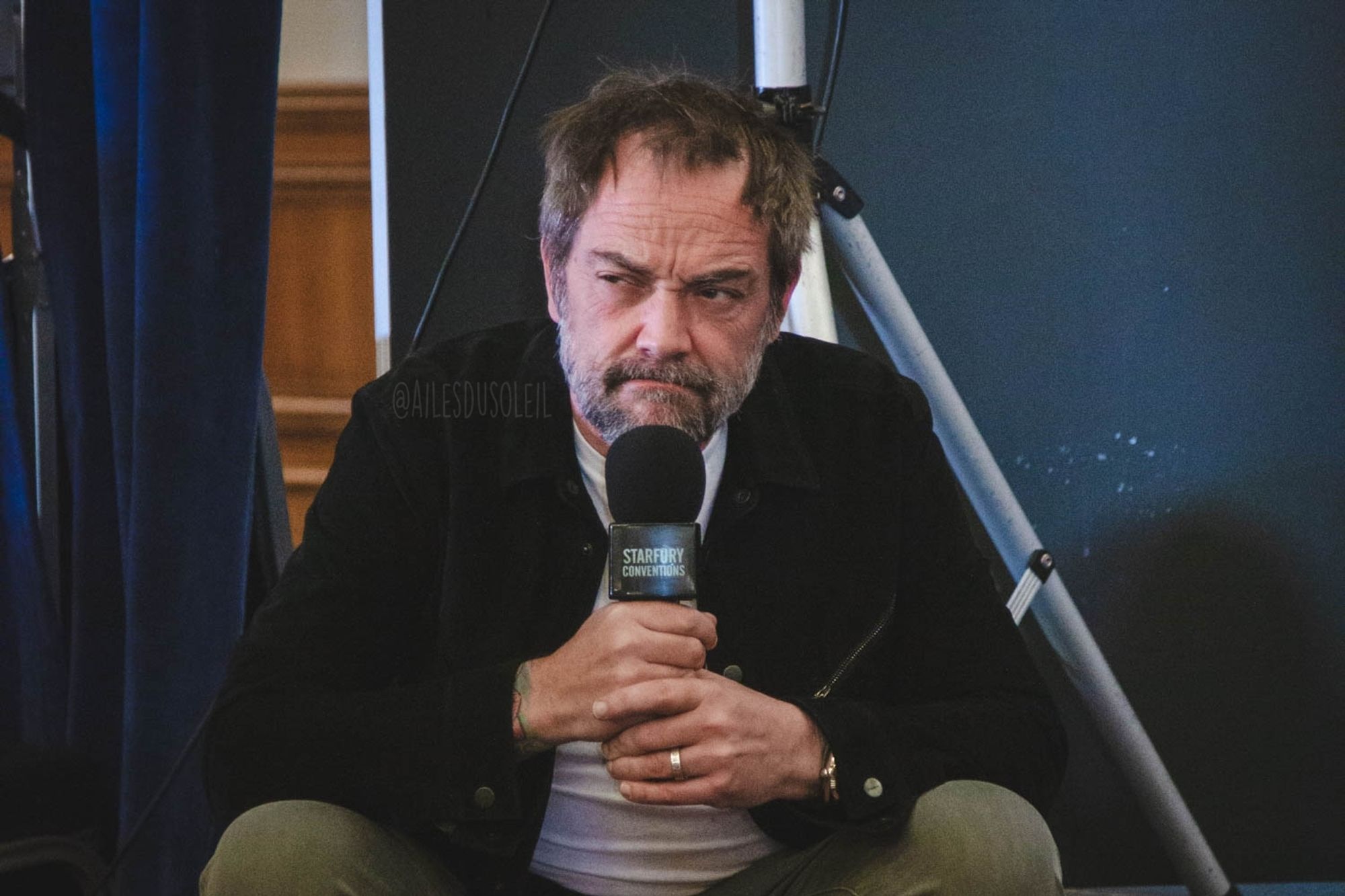 Mark is sitting on the edge of the stage, feet on the ground. He is facing the direction of the camera which is in the middle right of the stage. The stage is low so his knees are higher than his hips, spread a bit wider than his shoulders. He is leaning on his legs with his elbows, both holding up a microphone chin height. His face is grumpy - mouths sides turned down and eyebrows in a frown. His eyes are looking to the left of the photo. He is wearing gray pants, a white shirt and black jacket. His hair is messy and both hair and beard have white in it.