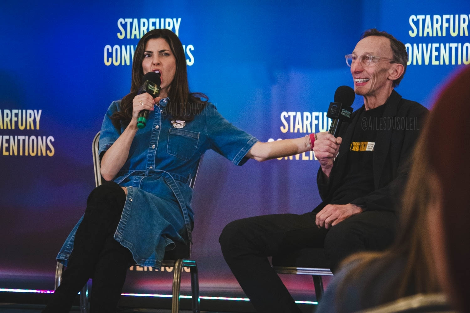 Cindy and Julian are sitting on chairs on stage facing the crowd to the left of the camera, about ⅔. Cindy is talking into her microphone she has in her left hand while her left hand is on Julians upper arm. She is making a face, mouth open like shouting and eyebrows scrunched. Julian is smiling into his microphone he has in his left hand mouth height. His right hand is on his lap. Cindy is wearing a long jean material button up dress that is blue and black made out of soft material over the knee boots. Her hair is dark brown and long. She has a ring on her right hand ring finger. Julian is wearing black jeans, black t-shirt and a black jacket. He has see-through plastic glasses on.