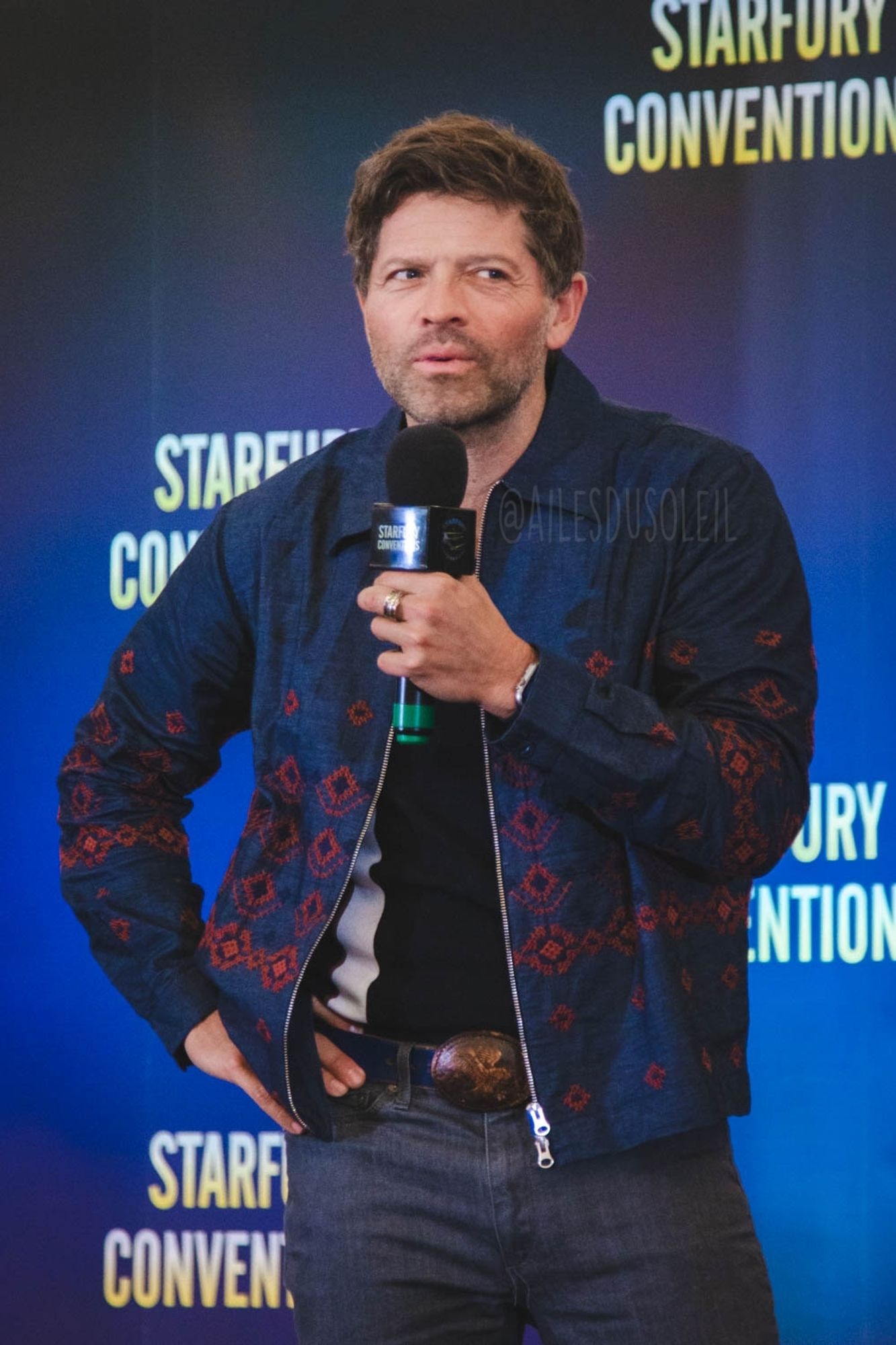 isha is standing on stage, turned a bit to the left of the camera towards the crowd, right hand on his hip and looking to the right of the photo with his eyes. He is mid talking into the microphone which he is holding in his left hand bow chin height. He is wearing dark gray jeans, a dark blue belt with a brown belt buckle, black polo shirt with a white stripe and a dark blue zip up jacket that has dark red-ish brown patterns on it.