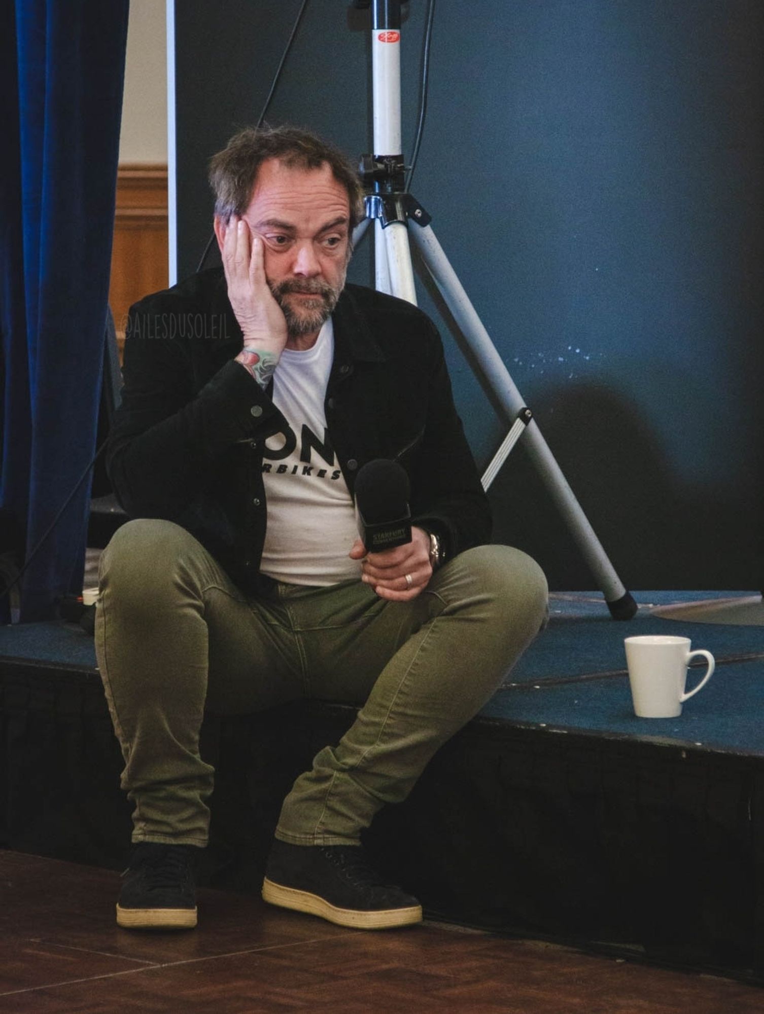 Mark is sitting on the edge of the stage, feet on the ground. He is facing the direction of the camera which is in the middle right of the stage, but body is slightly turned to the right of the photo. The stage is low so his knees are higher than his hips, spread a bit wider than his shoulders. He is leaning on his legs with his elbows. The right hand has his head resting on it while the left hand is holding the microphone up. He is staring into space in front and a bit to the right of him. He is wearing dark blue shoes, gray pants, a white shirt and black jacket. His hair is messy and both hair and beard have white in it. There is a white cup of most likely tea next to him on stage.