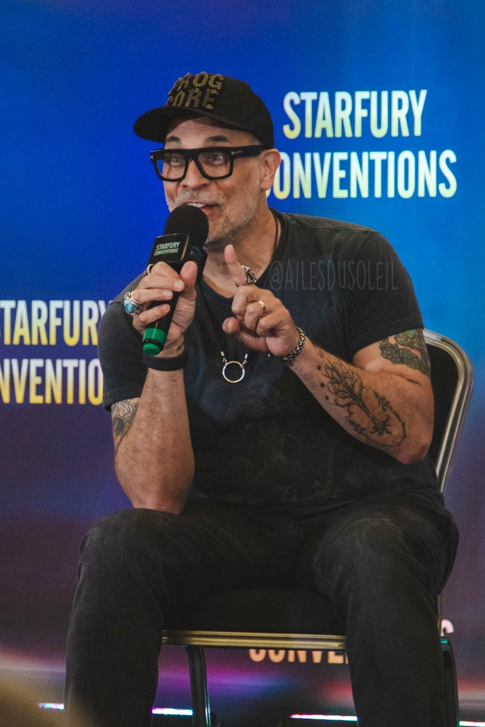 Todd is sitting on a chair on stage. He is turned about ⅔ towards the left of the camera, facing the crowd. He is leaning slightly forward on right elbow, which is on his right thigh. He is talking into the microphone he has in his right hand. He looks excited, eyes open and alert, talking into the microphone, slightly smiling. His left arm is bent and closer to his body, but his forearm is leaning forward, pointer finger up for explaining. He is wearing black jeans, black t-shirt and a black baseball cap. He has black framed glasses, some tattoos, wears multiple rings and a ring shaped necklace.