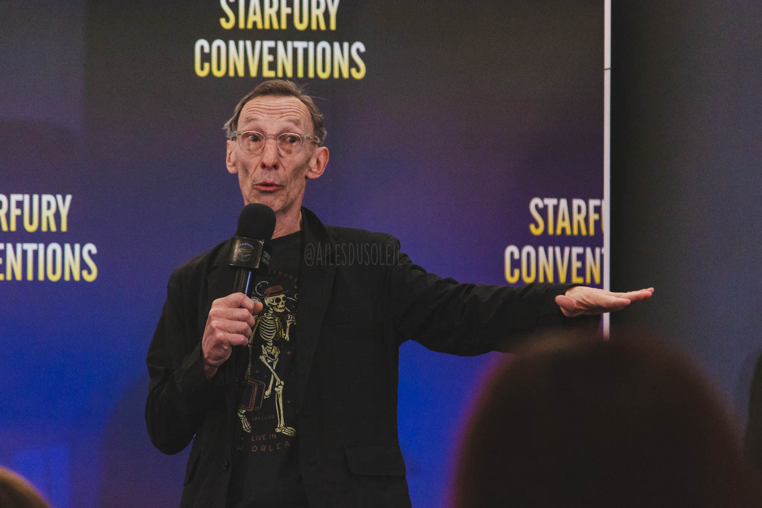 Julian is standing on stage, visible belt up and turned towards the crowd slightly to the left of the camera. He is looking to the right of the camera.. He is speaking into his microphone which he is holding with his right hand to his mouth. His left arm is stretched out to his side a bit mid motion explaining something. He is wearing a black t-shirt with a skeleton on it and a black blazer. He has see-through rimmed glasses.