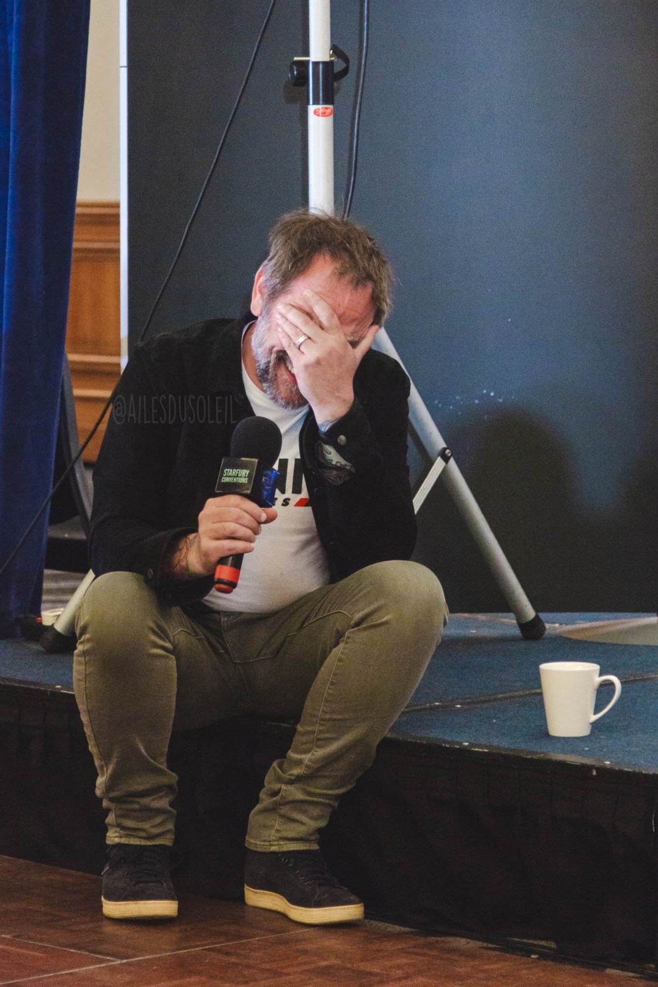 Mark is sitting on the edge of the stage, feet on the ground. He is facing the direction of the camera which is in the middle right of the stage. The stage is low so his knees are higher than his hips, spread a bit wider than his shoulders. He has his left hand on his face while laughing, the elbow is leaning on his left leg. The microphone is in his right arm that is leaning on his right leg. He is wearing gray pants, a white shirt and black jacket. His hair is messy and both hair and beard have white in it.