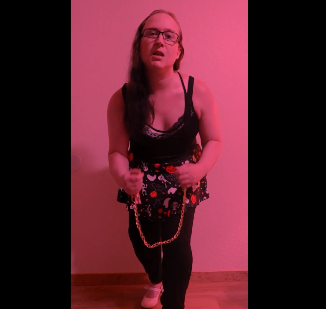 image is tinted red. me in a black outfit with a halloween themed skirt over my leggings. the skirt has pumpkins and ghosts on it and is very festive and spooky. holding a leash with a gold colored chain and pink handle. white kitten heels adorn my feet and my hair is long and black and i am a white trans woman.