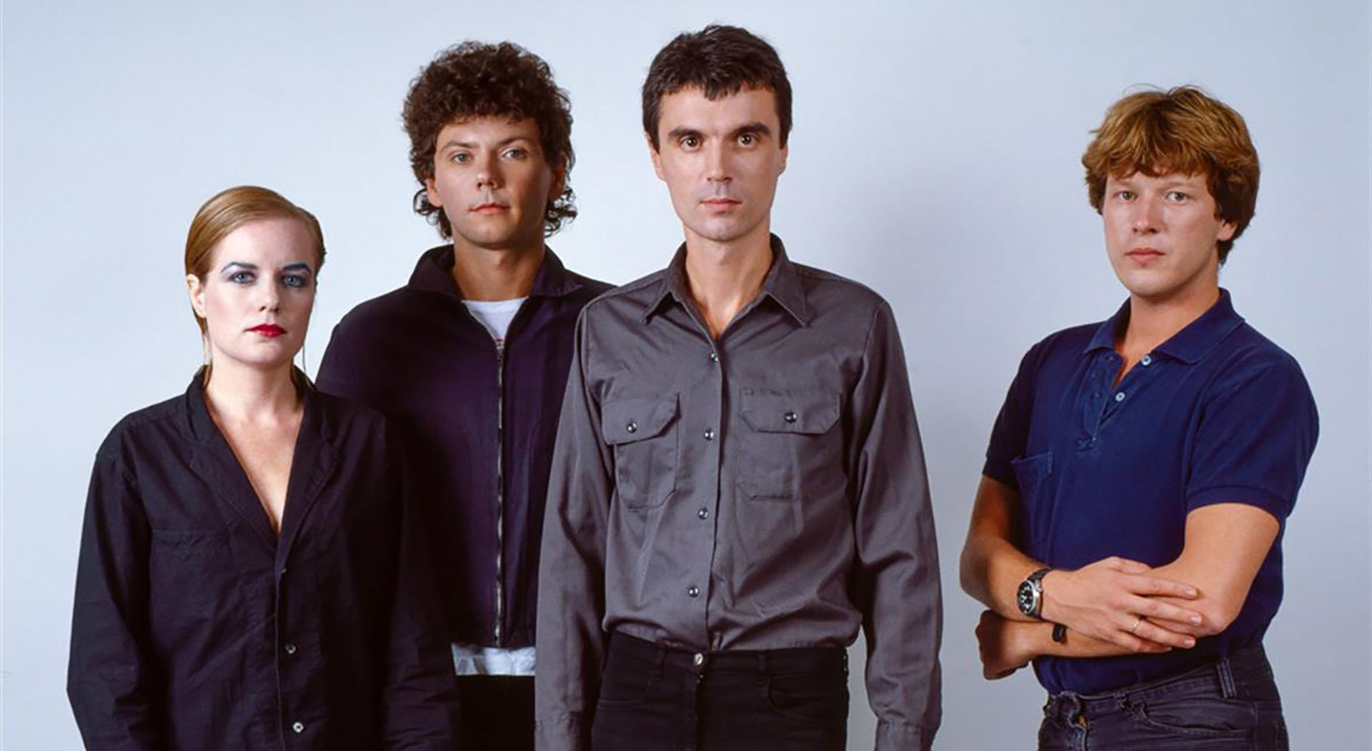  David Byrne in a grey shirt and black pants short black hair  Jerry Harrison in a black shirt frizzy black mullet black pants  Tina Weymouth in a black shirt red lipstick blue eyeliner brown hair Chris Frantz in a blue shirt black pants short brown hair folding arms together. hot