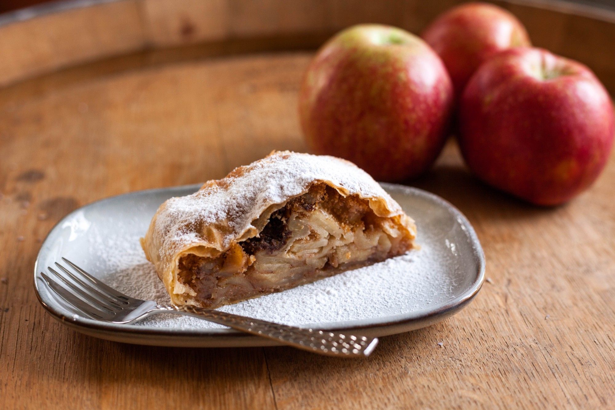 Im Vordergrund ein Stück Apfelstrudel mit selbstgezogenem Teig auf einem hellgrauen Dessertteller und einer Gabel. Im Hintergrund drei rote Äpfel aus dem Garten einer Freundin. Alles zusammen steht auf einem alten Mörwald Weinfass aus Holz.