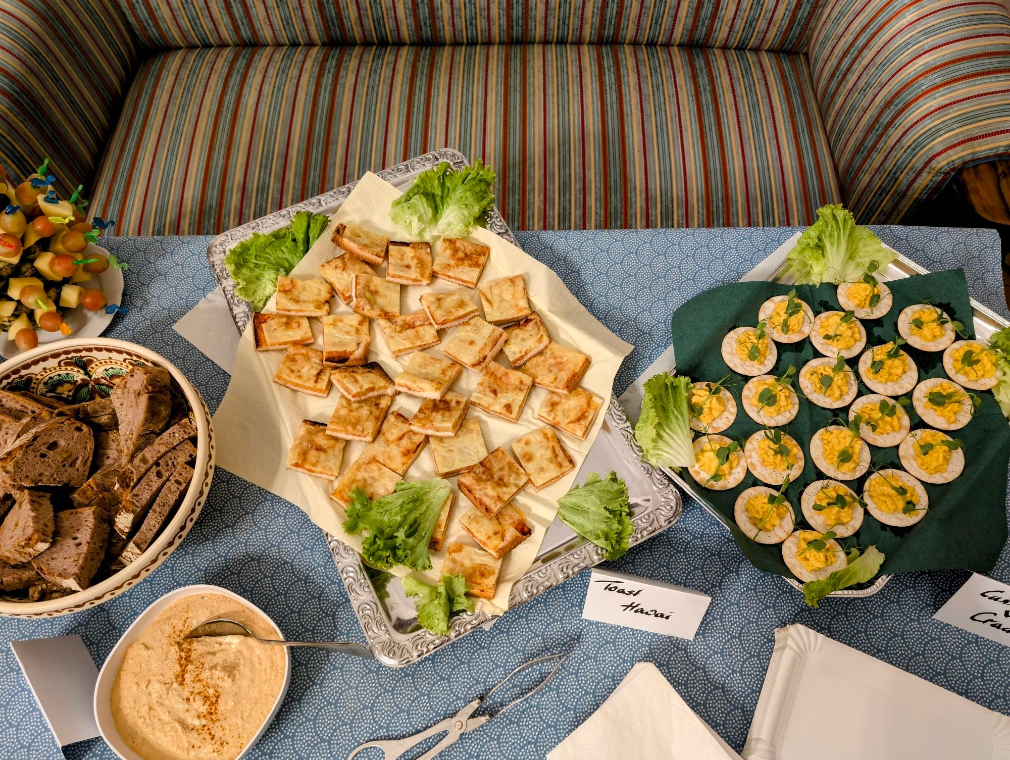 Auf einem Tisch. Mit blauer Tischdecke: Walnussbrot aus eigener Produktion und Liptauer, Toast Hawaii 2.0, Cracker mit Käse-Curry-Creme und Erbsensprossen. Im Hintergrund eine alte Couch in der ÖGFL, Sitzgrund für viele Autorinnen und Autoren seit den 60er Jahren.