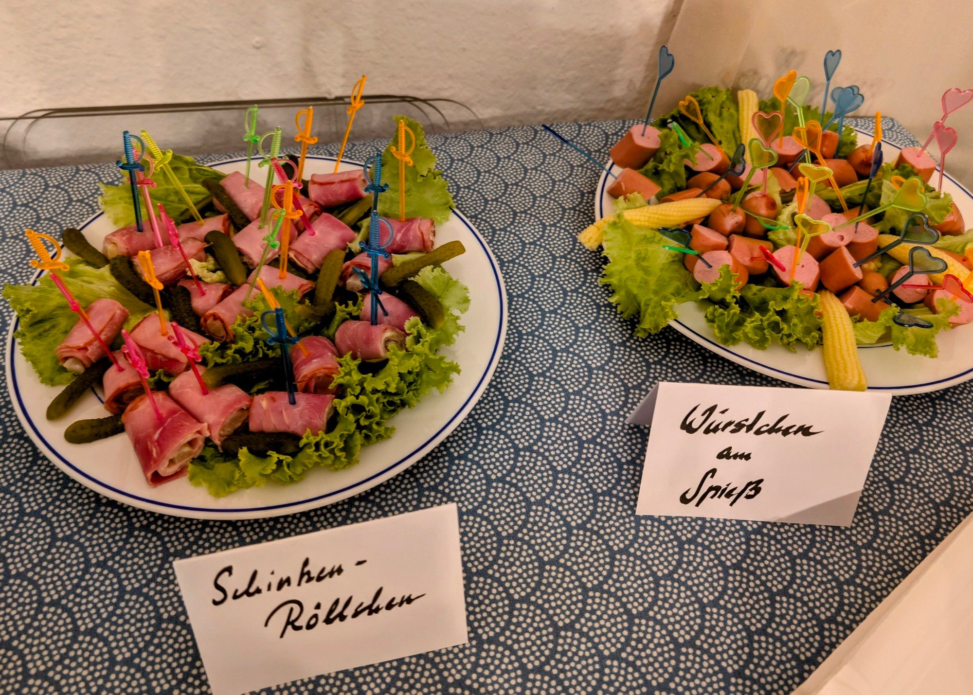 Zwei Teller, einer mit Spargel-Schinkem-Röllchen, Cornichos und Salat als Verzierung. Auf dem Teller rechts statt den Cornichos Mini Maiskolben und statt den Röllchen Frankfurter Würstl Stücke. Aufgespießt auf beiden Tellern mit bunten Plastikspießen.