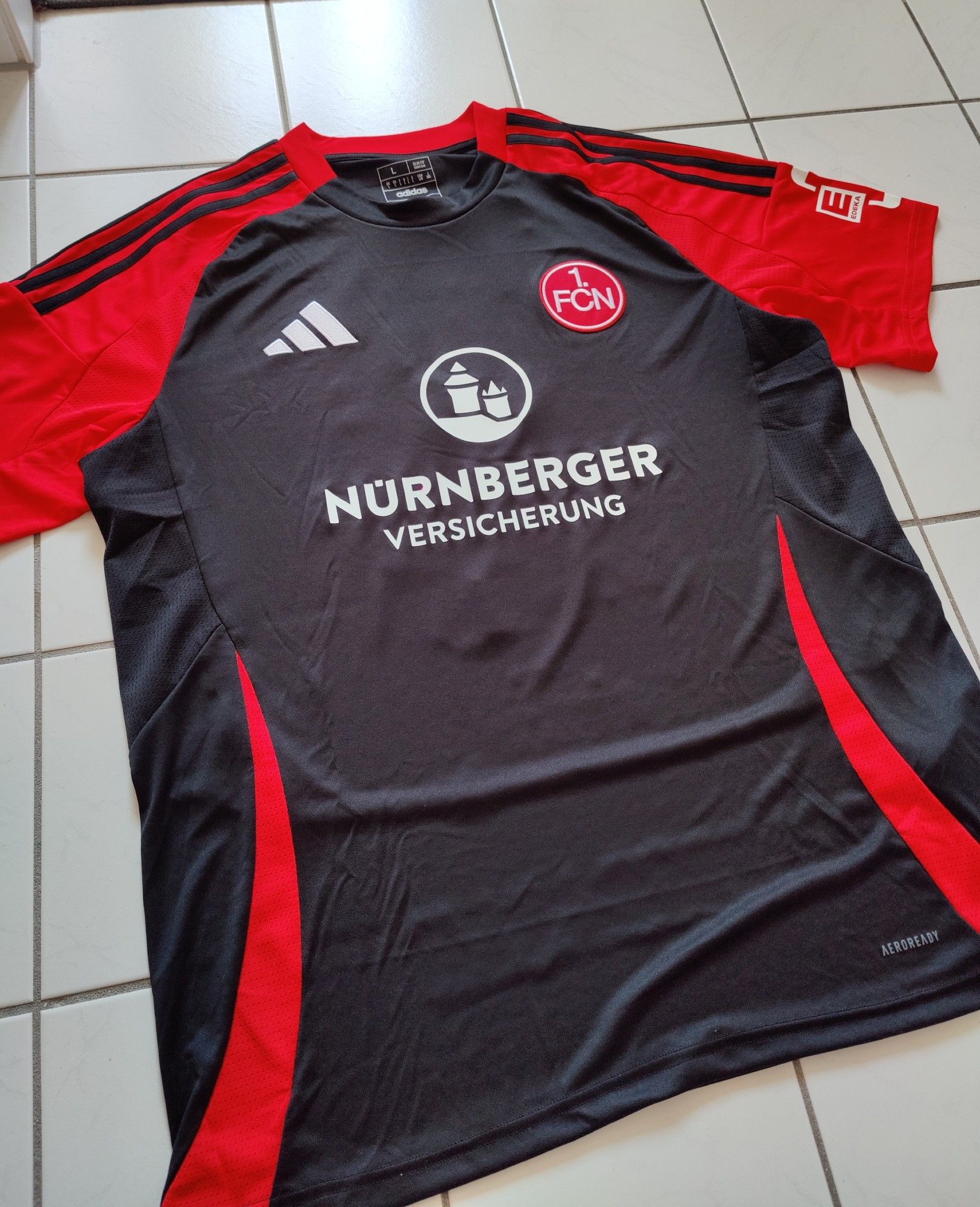 Das dritte Trikot der Clubfrauen der Saison 2024/25, auf einem weißen Fließboden liegend. Das Trikot ist überwiegend schwarz mit knallig roten Akzenten, wie die Streifen vorne, die sich in einem Bogen jeweils nach außen auf beiden Seiten vom unteren Saum bis knapp unter der Brust hochziehen, dabei immer schmaler werden und spitz enden. Die Ärmel, als auch die Schultern, sind in dem selben Rot gehalten, die typischen Adidas-Streifen auf der Schulter in schwarz. Das FCN-Wappen ist unverändert, das Logo von Adidas und der Nürnberger Versicherung sind weiß.