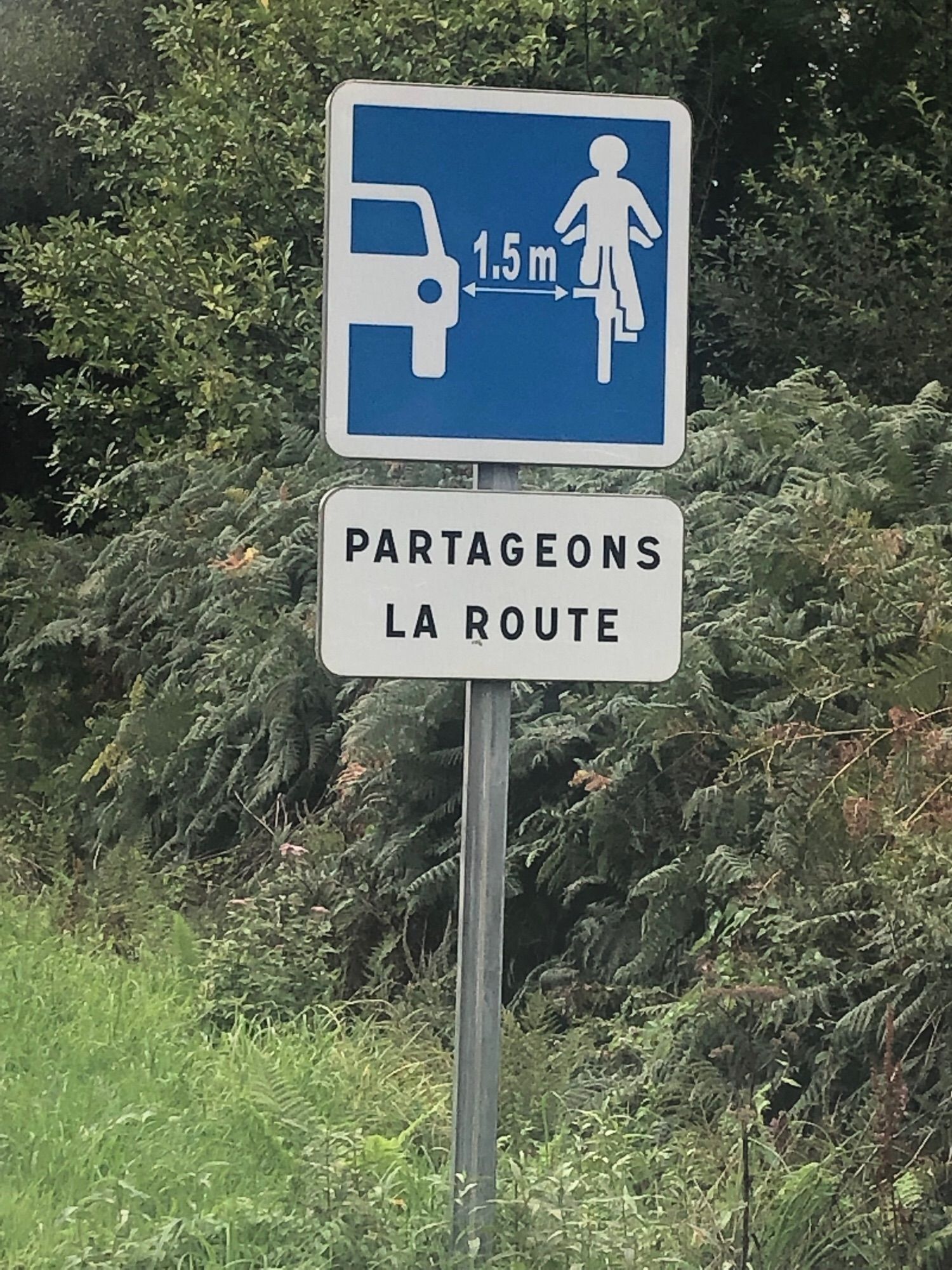 Hinweisschild (blau) am Straßenrand, das mit Piktogramm auf den Abstand von Pkw- zu Fahrradverkehr (1,5 m) verweist. Ein zweites (weißes) Schild besagt auf Französisch „Lasst uns die Straße teilen“ (Partageons la route). Am Straßenrand ist noch grünes Buschwerk und Gras zu sehen.