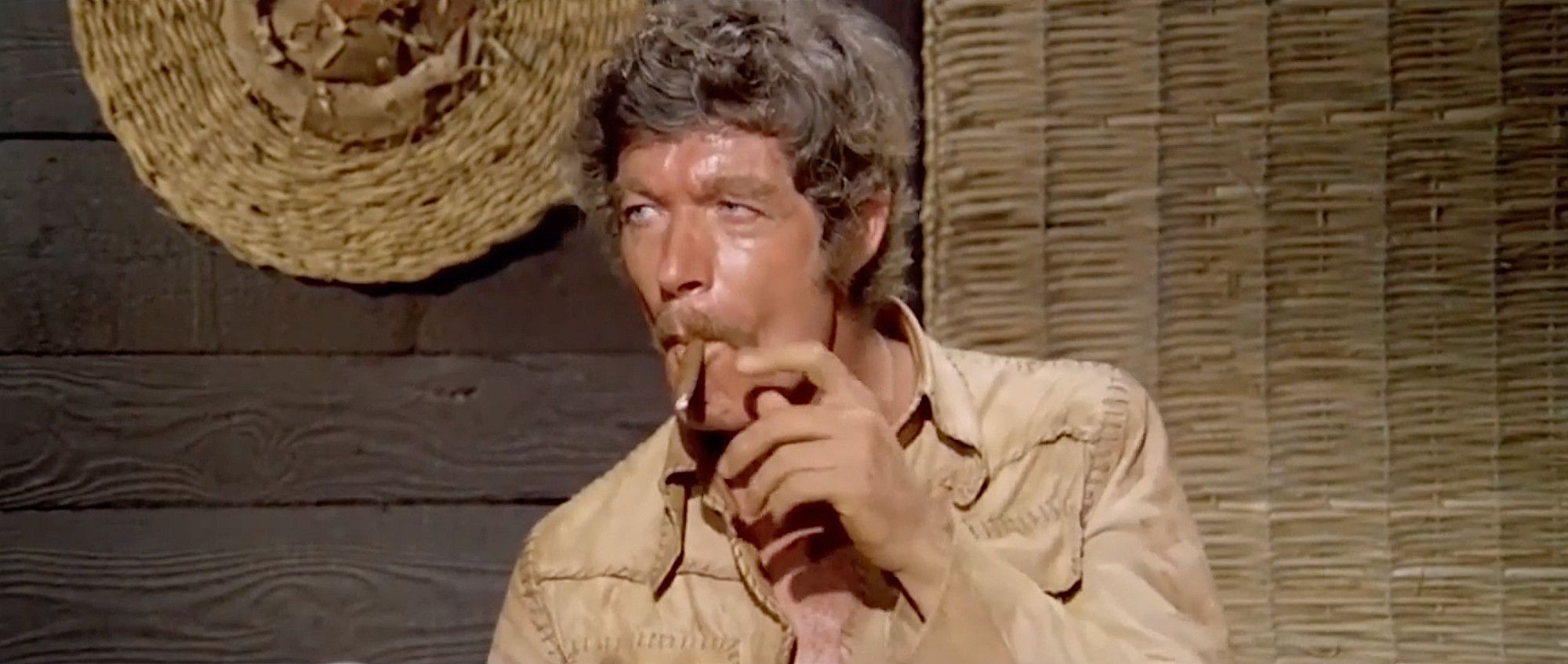 Stephen Boyd smoking a cigar, a straw hat and mat hanging on the wall