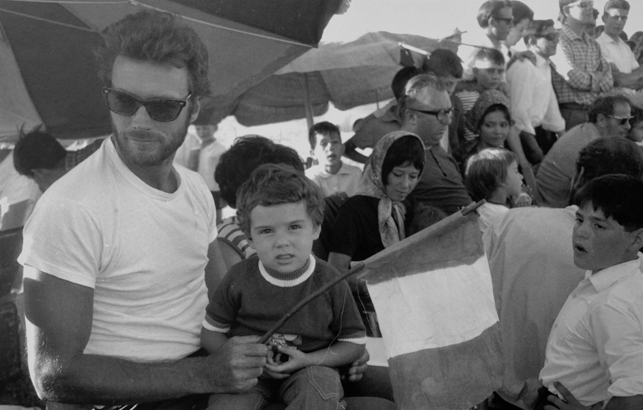 Clint Eastwood, Sergio Leone and Chelo Alonso sit among the villagers in the shade of umbrellas to watch a scene from the film being shot.