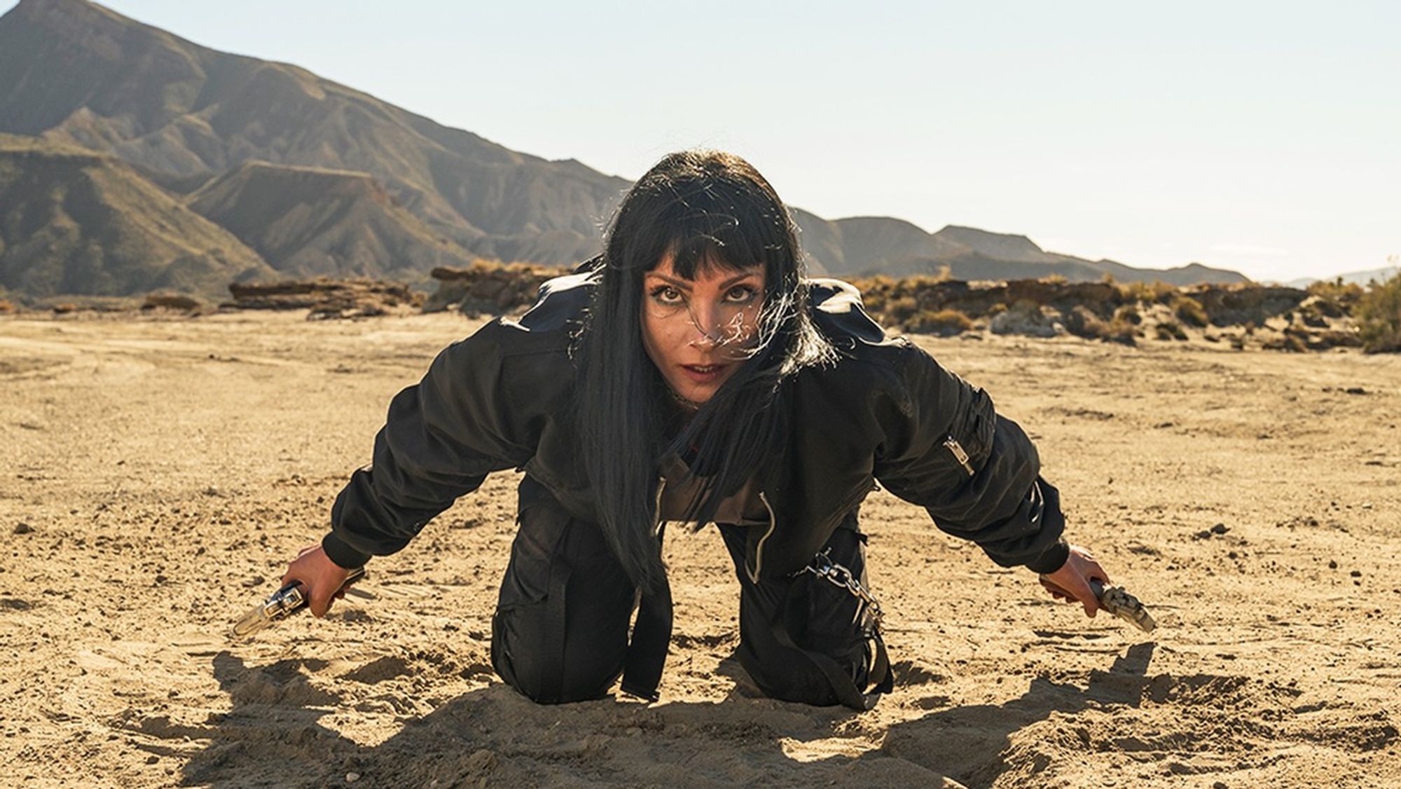 Najwa Nimri dressed in black with an intense look, kneeling in the desert with a revolver in each hand. Shot in La Salinillas.