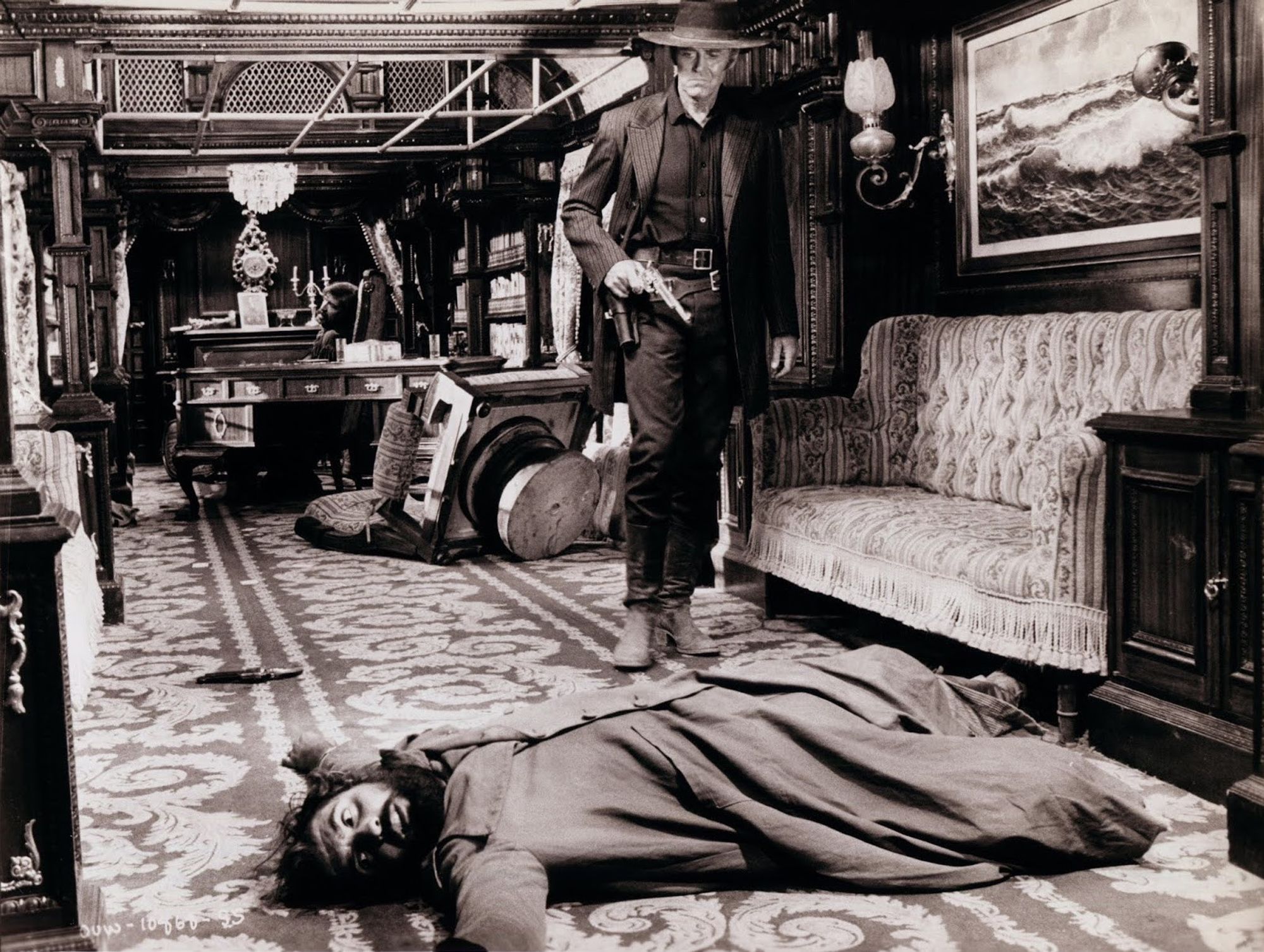 Man dressed in black, revolver in hand, cautiously advances into a richly furnished and decorated carriage. In front of him, the body of a gang member lies on the floor.